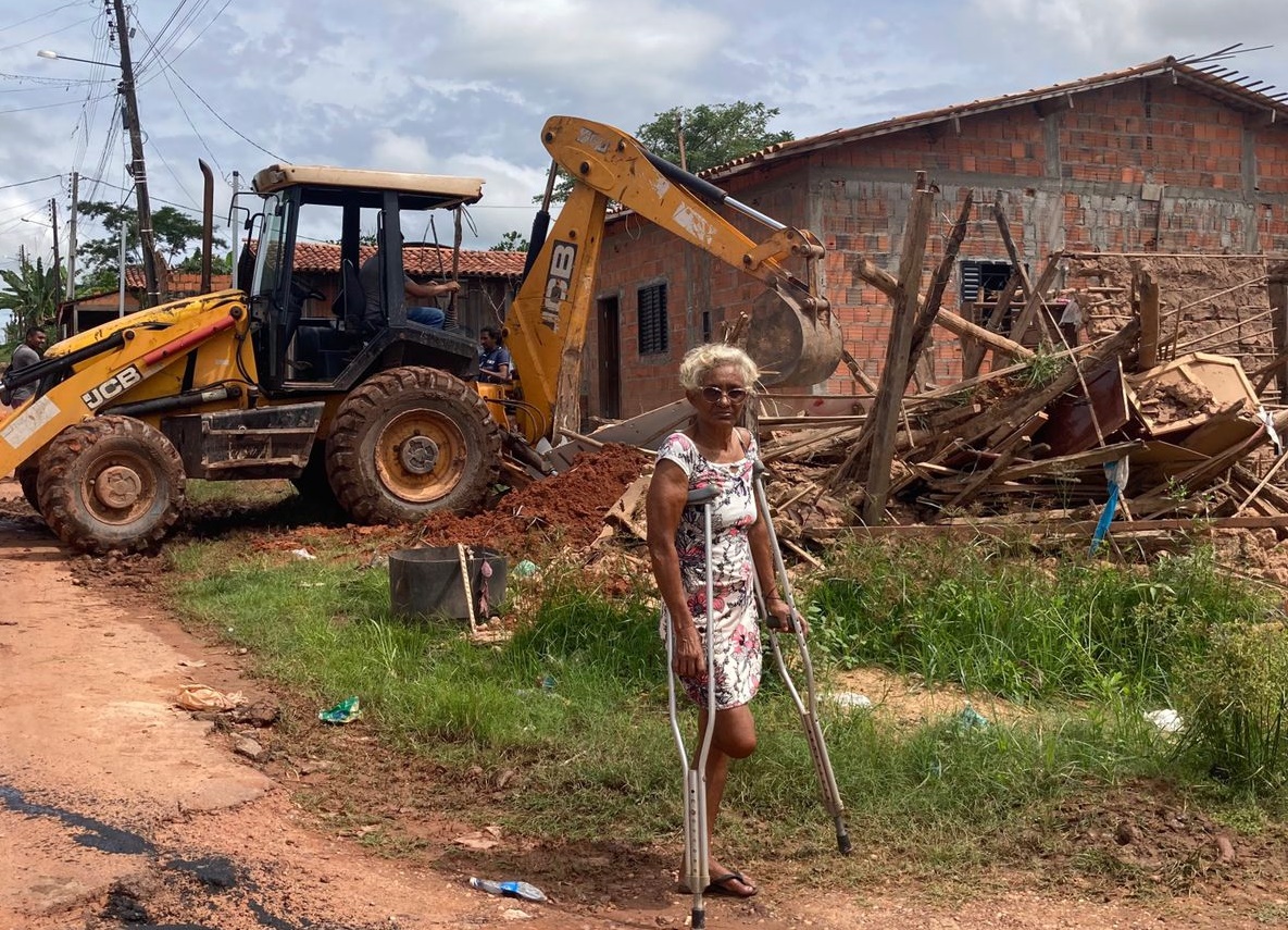 Mulher idosa, de vestido, com moleta, em frente à obra de construção de casa, com um trator na frente