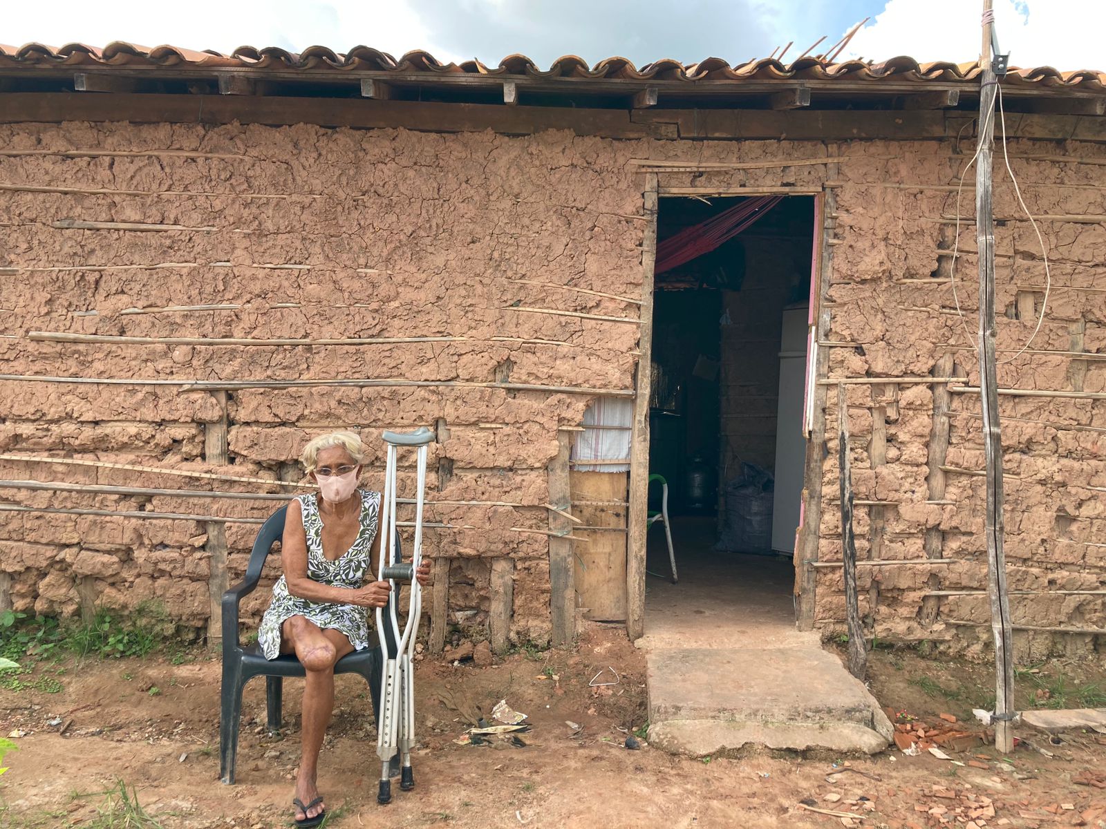 Mulher idosa, com moletas e perna amputada, em frente a casa de taipa.