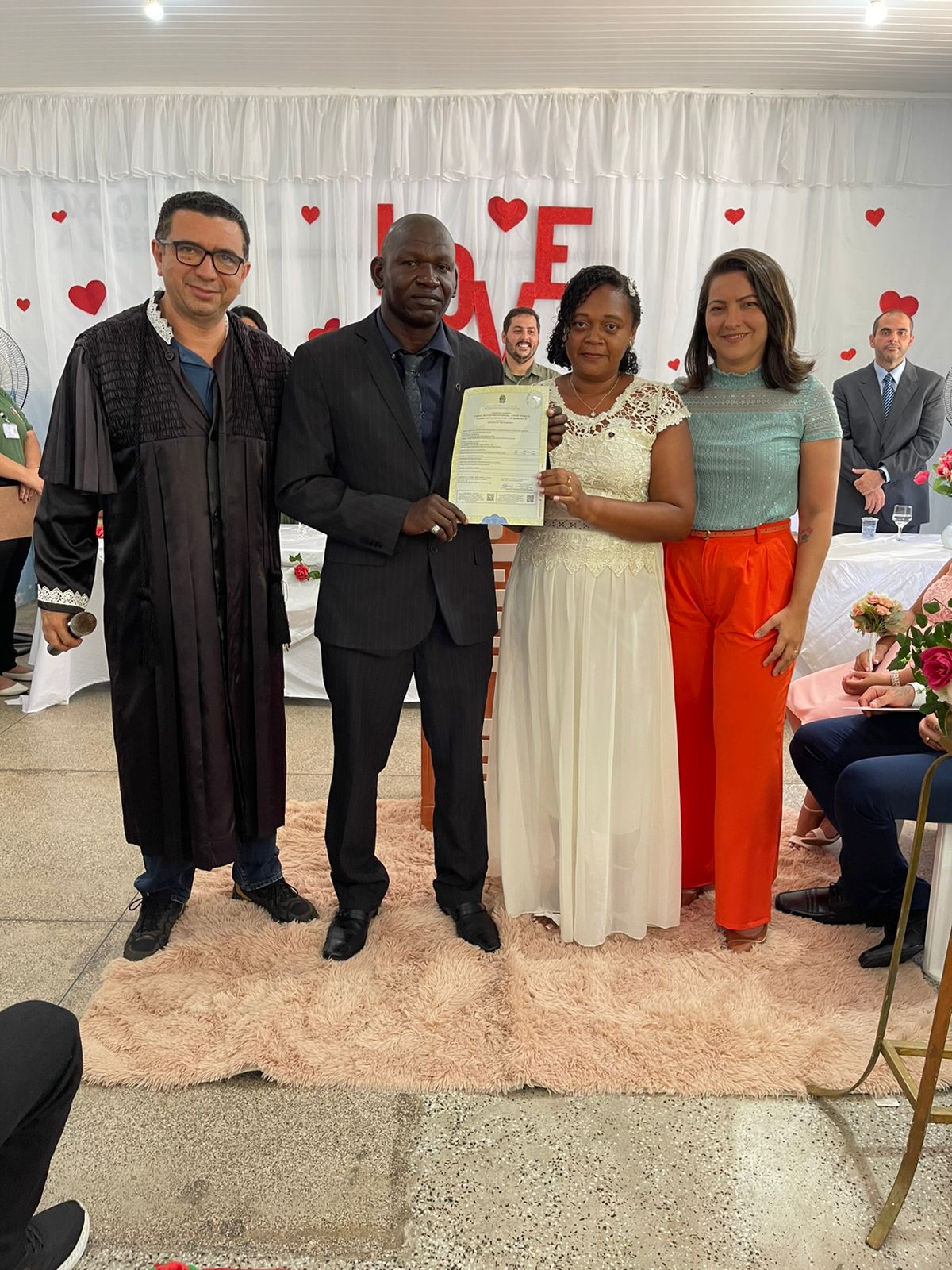 Homem negro, de terno preto, ao lado de mulher parda, de vestido de noiva, entre mulher de calça laranja e blusa verde e homem de toga preta.