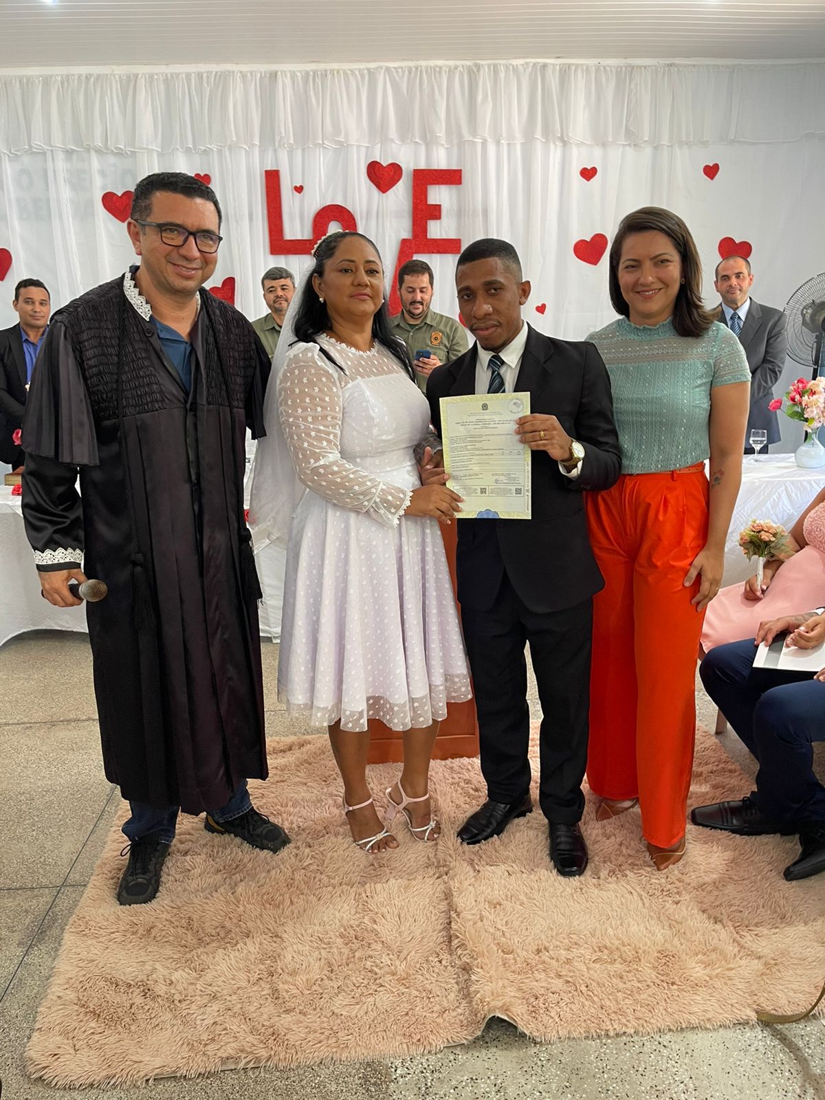 Homen negro, de terno preto, com mulher parda, de roupa de noiva, entre um juiz de toga preta e mulher de calça laranja e blusa verde.