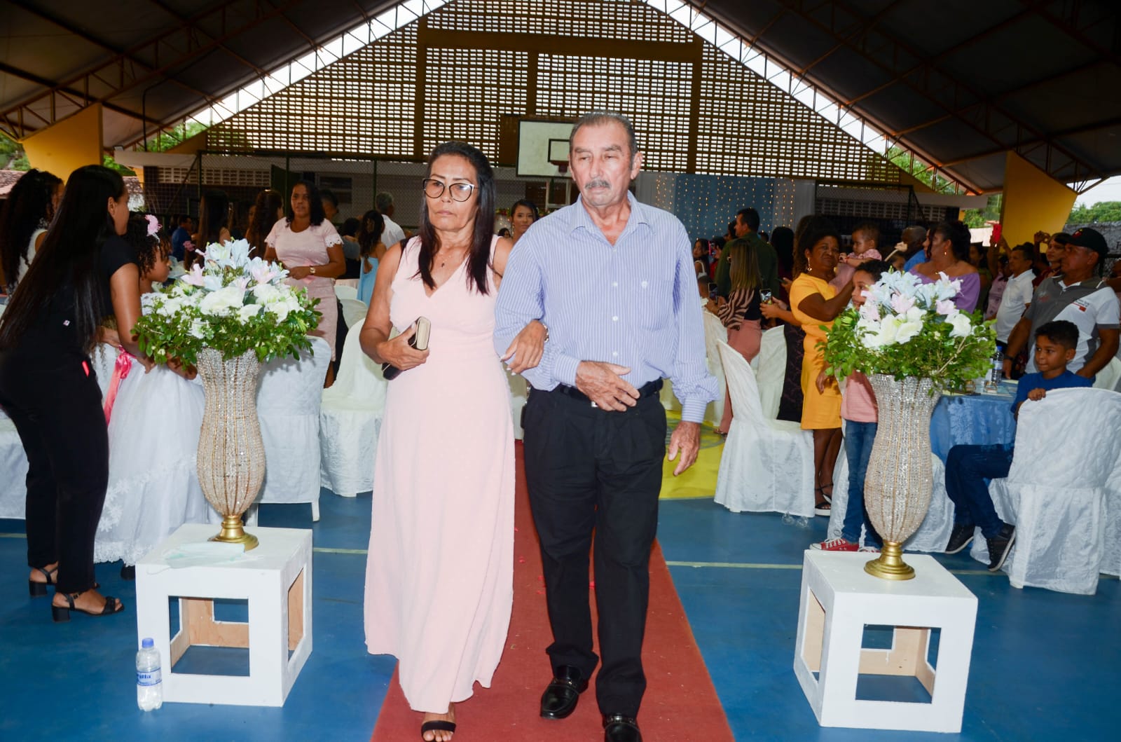 Casal de homem e mulher idosos, ela de branco, ele de camisa azul e calça preta, de pé entre dois vasos de flores, ao lado de casais sentados em cerimônia de casamento.