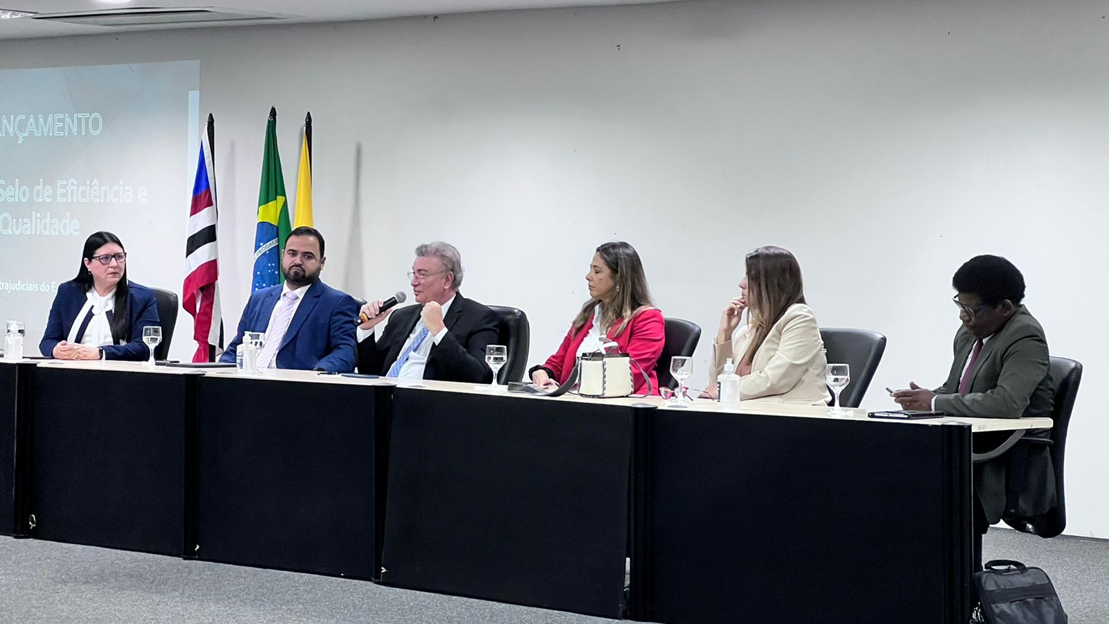 Três homens de terno escuro e três mulheres em trajes formais, em mesa diretora de evento, ao lado das bandeiras do Brasil, do Maranhão e do Município de São Luís. Em salão de paredes brancas.