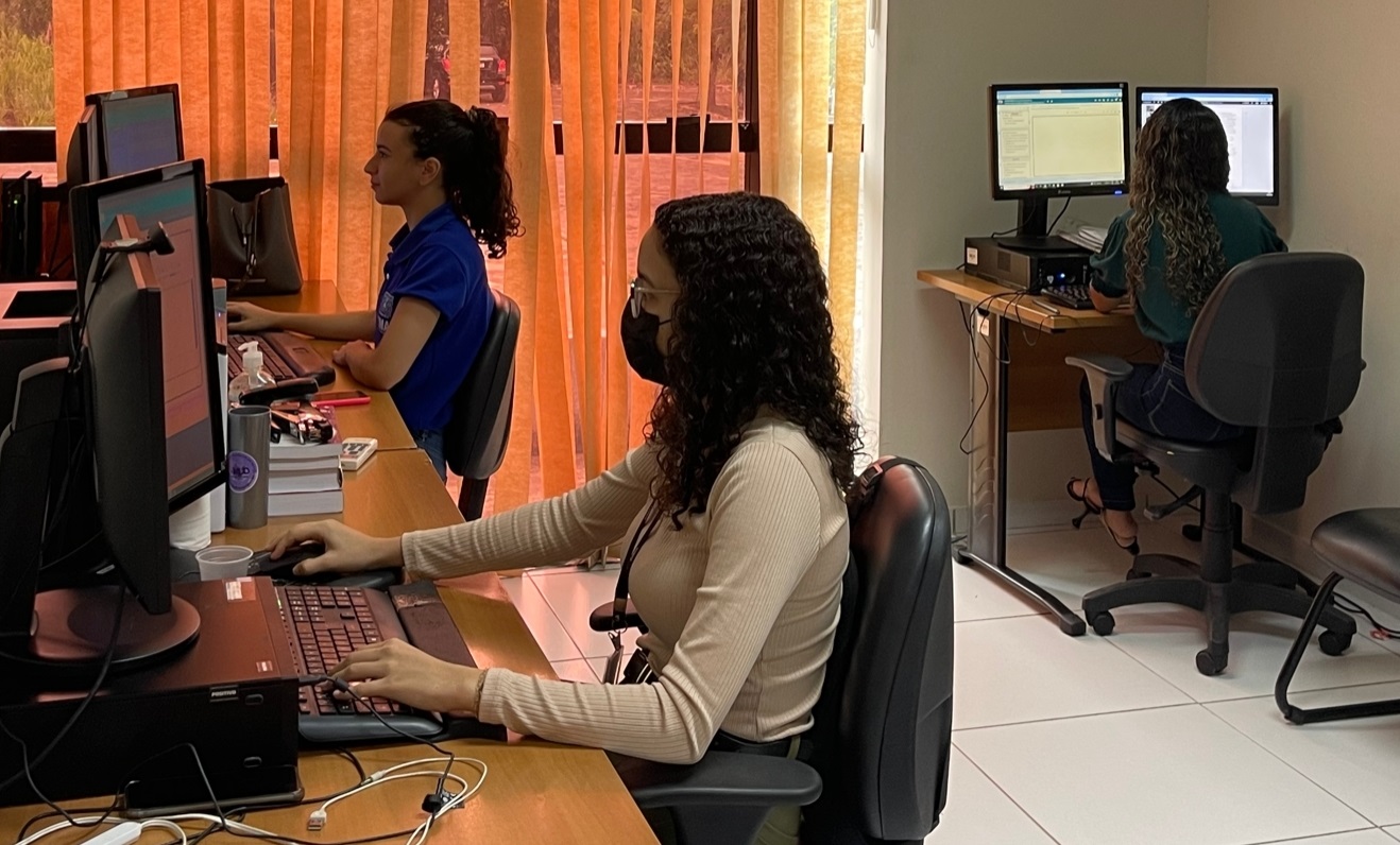 TrÊs mulheres sentadas diante de computadores, em sala de trabalho, tendo ao fundo uma vidraça com cortina persiana bege clara. Duas à esquerda e uma à direita., no canto.