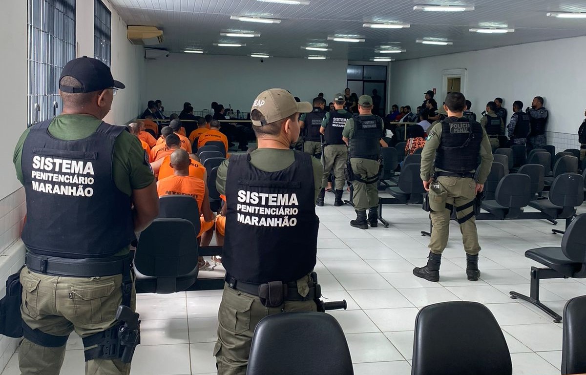 Foto de pessoas sentadas em auditório de audiência na Justiça, de costas. Em primeiro plano, policiais militares de costas e de pé.