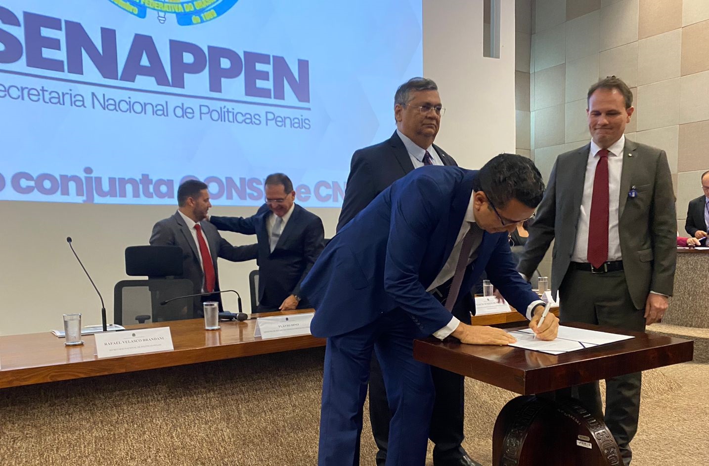 hOMEM BRANCO,C ABELOS PETOS, TERNOA ZUL, ASSINA DOCUMENTO SOBRE MESA, AO ALDO DE HOMEM BRANCO, TERNO VERDE, DE PÉ, E DIANTE DE HOMEM PARDO, TERNO ESCURO, DE PÉ.AO FUNDO, PAINEL NA PAREDE COM NOME SENAPPEN, EM AUDITÓRIO.