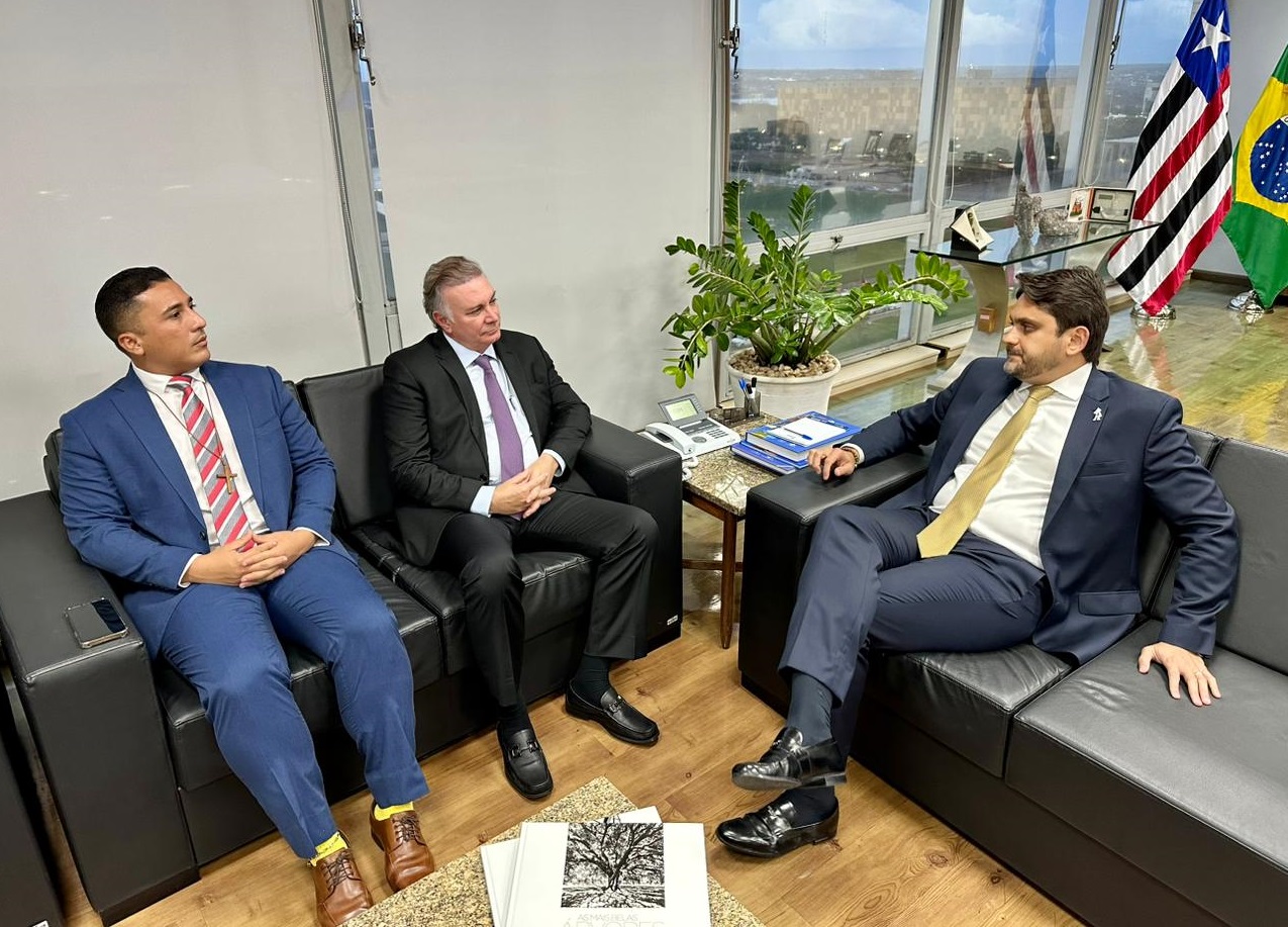 Três homens, de terno, um à direita e dois à esquerda, sentados em sofá preto, em sala de trabalho, com bandeiras do Brasil e do Maranhão ao fundo.