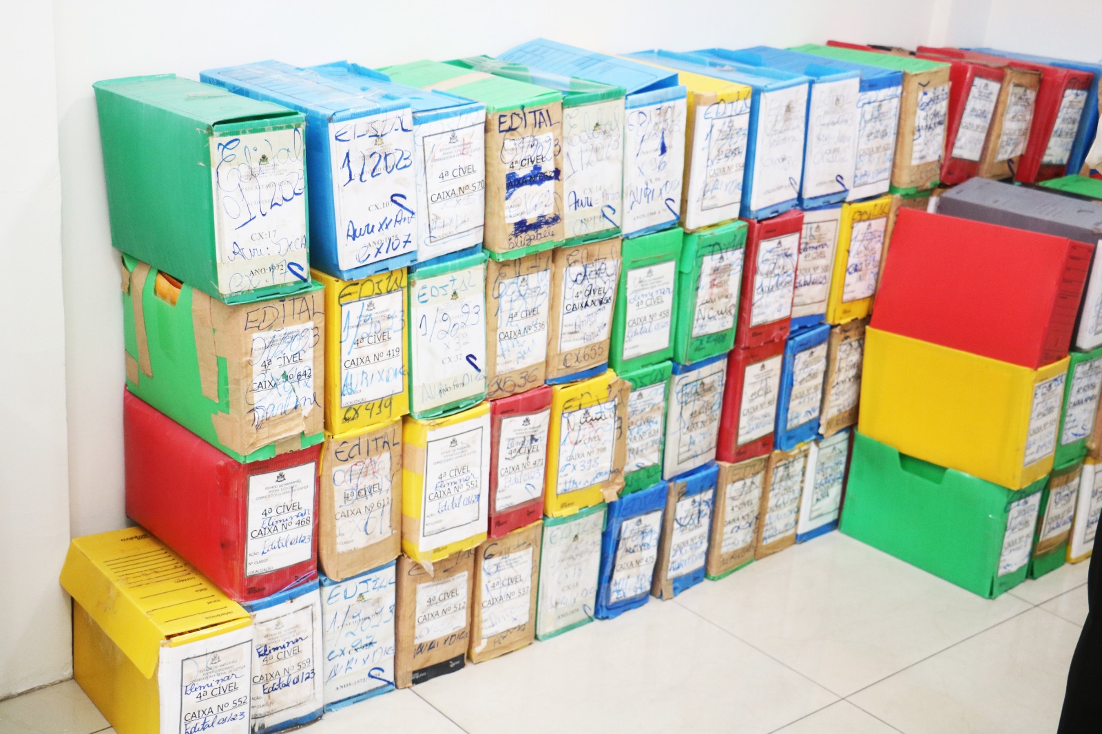 quatro pilhas de caixas de plástico coloridas contendo processos judiciais encostadas em parede branca e sobre piso branco