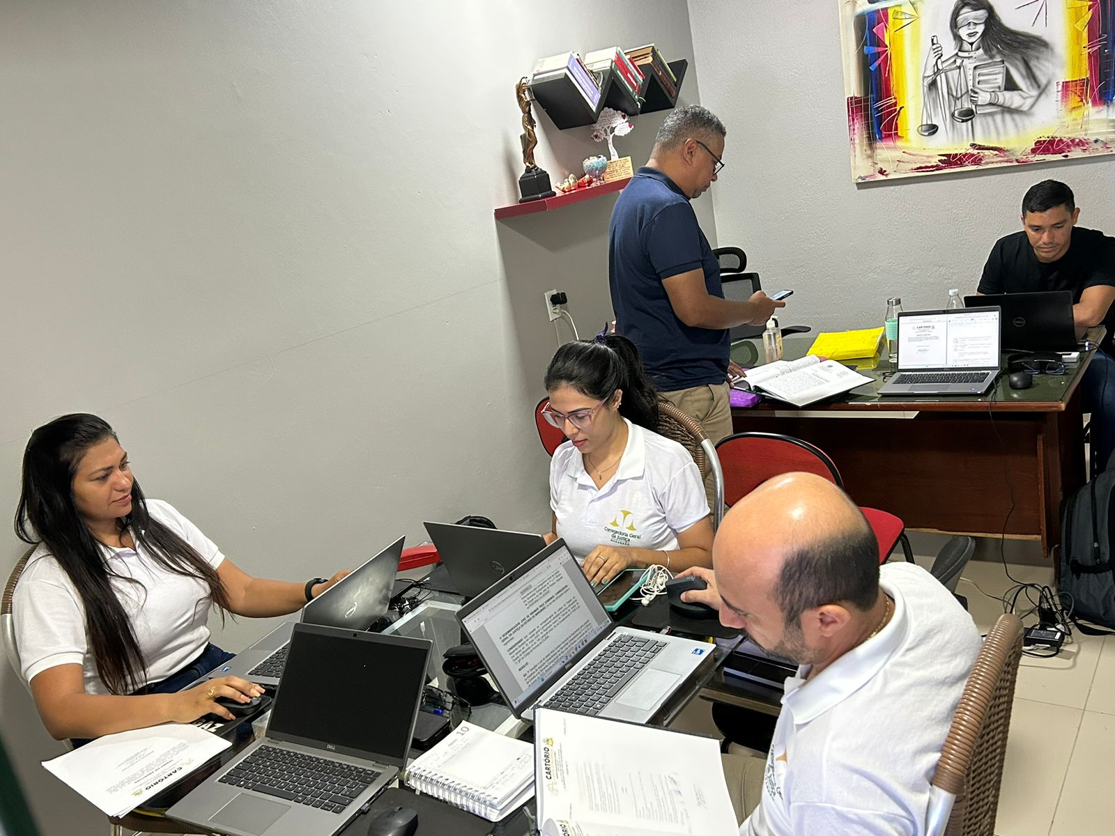 Foto horizontal mostra homens e mulheres de camisa branca, sentados diante de computador, em mesa de trabalho.