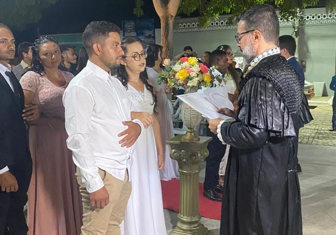 na imagem, um juiz, de toga preta, realizando o casamento de alguns casais em fila, em pé, numa praça