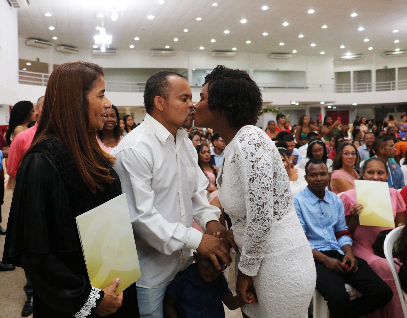 Foto horizontal de mulher parda, sorri com pasta de papel na mão esquerda, para casal de homem e mulher negros, que se beijam. Ao fundo, pessoas sentadas em auditório.