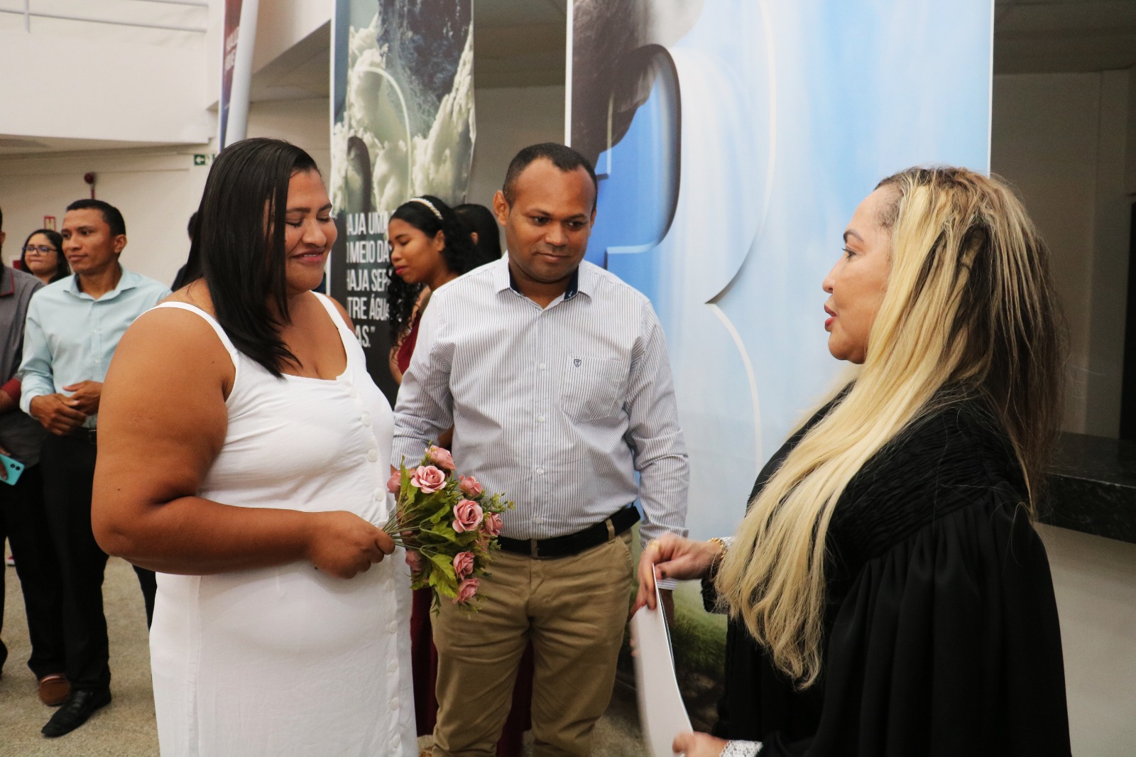 Casal de noiva parda de vestido branco e home pardo de camisa azul e calça befe, tendo ao lado mulher de cabelos louros e toga preta.