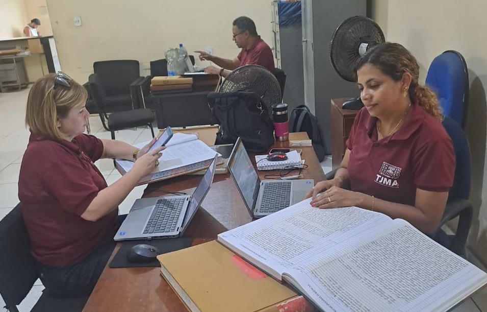 Foto horizontal de sala de cartório, com duas mulheres de camiseta cor de vinho analisando livros diante de computador. Ao fundo homem pardo sentado.