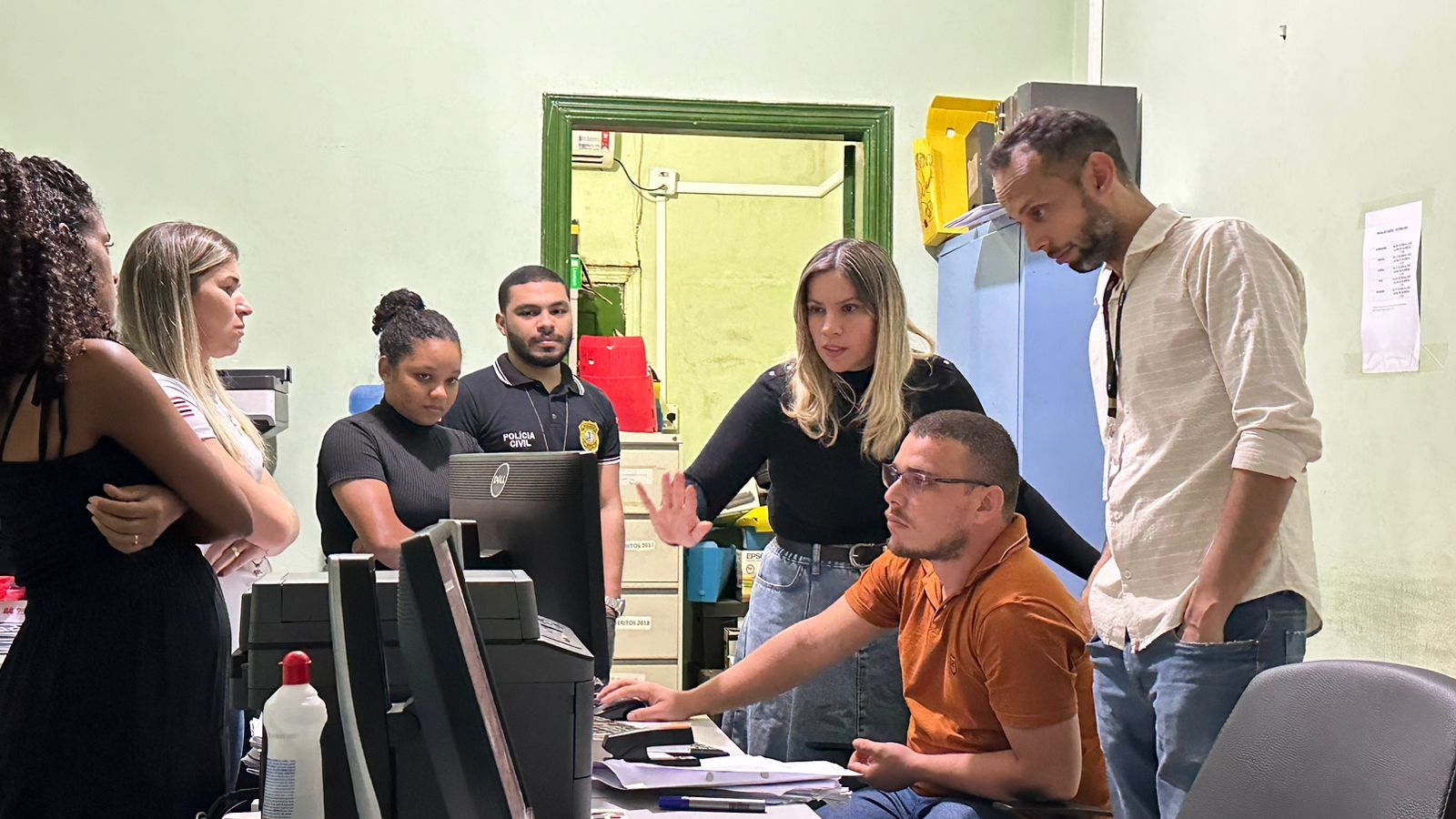 FOTO HORIZONTAL, COLORIDA, DE DOIS HOMENS - UM SENTADO - E UMA MULHER OLHANDO PARA COMPUTADOR SOBRE MESA EM SALA DE TRABALHO, OBSERVADO POR OUTRAS PESSOAS, DO LADO ESQUERDO DA FOTO.