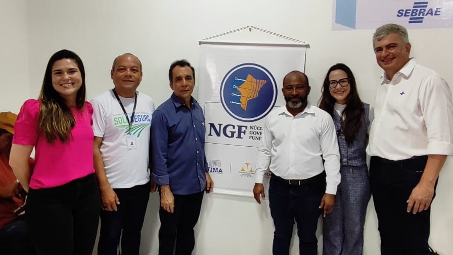 FOTO HORIZONTAL, COLORIDA, DE DUAS MULHERES E QUATRO HOMENS DE PÉ, DIANTE DE PAREDE CLARA, COM CARTAZ PENDURADO COM NOME NGF E FIOGURA CIRCULAR AZUL COM MAPA DO MARANHÃO NA COR AMARELA.