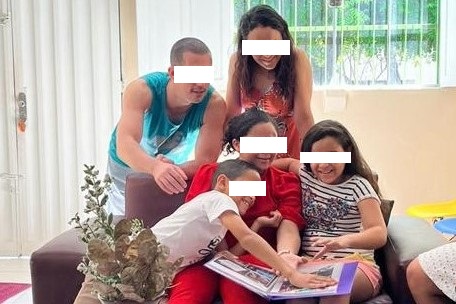 FOTO HORIZONTAL DE FAMÍLIA EM CASA. DUAS MENINAS E UM MENINO SENTADOS EM SOFÁ VERMELHO OLHAM ÁLBUM DE FOTOS. CASAL, DE PÉ, ATRÁS DO SOFÁ. AO FUNDO, PAREDE EM CLOR CALA COM PORTA E JANELA.