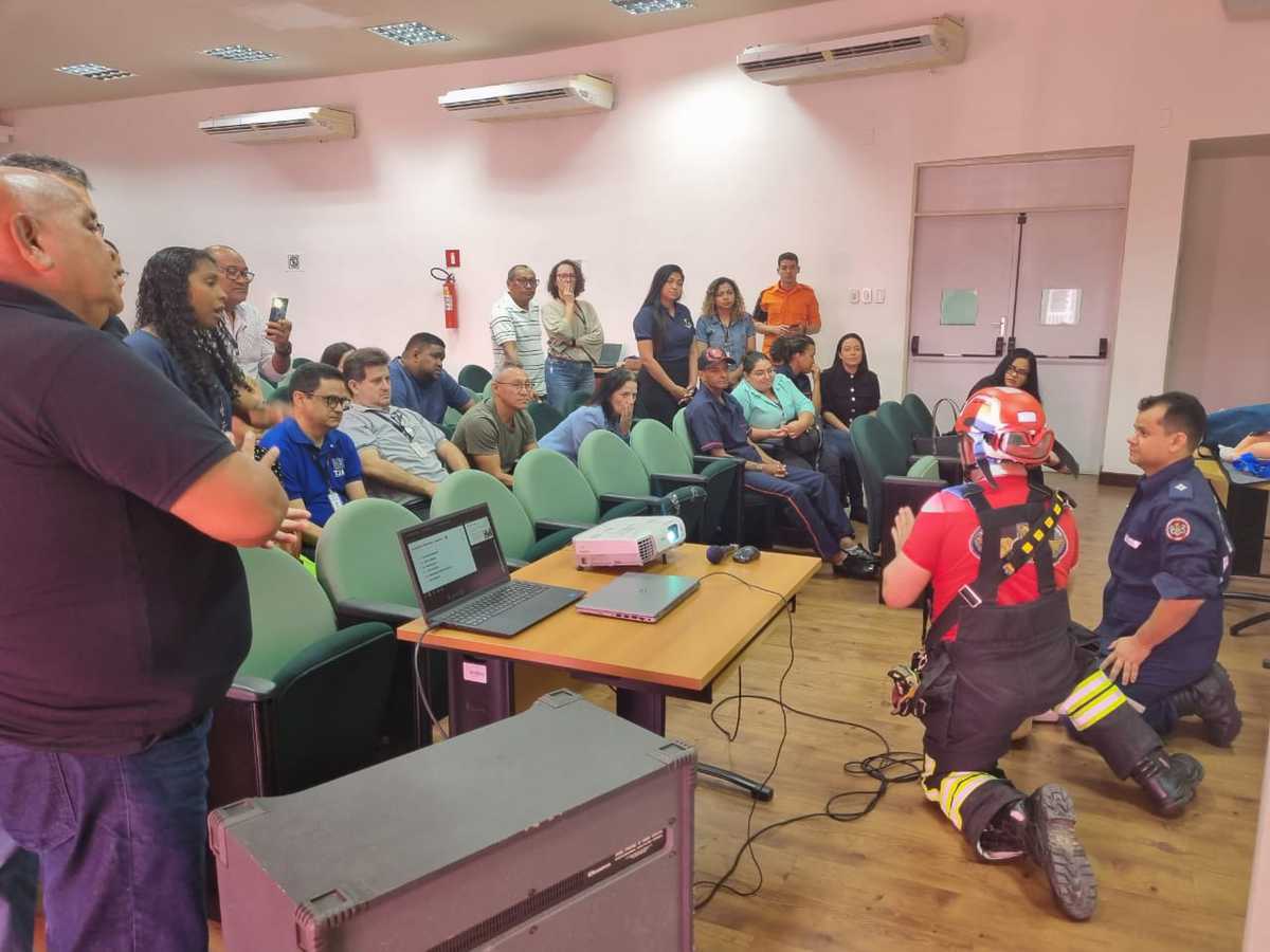 FOTO HORIZONTAL, COLORIDA, DE DOIS MILITARES AGACHADOS JUNTO A BONECO, DIANTE DE HOMENS E MULHERES SENTADOS E DE PÉ EM AUDITÓRIO, DURANTE CURSO DE PRIMEIROS SOCORROS.
