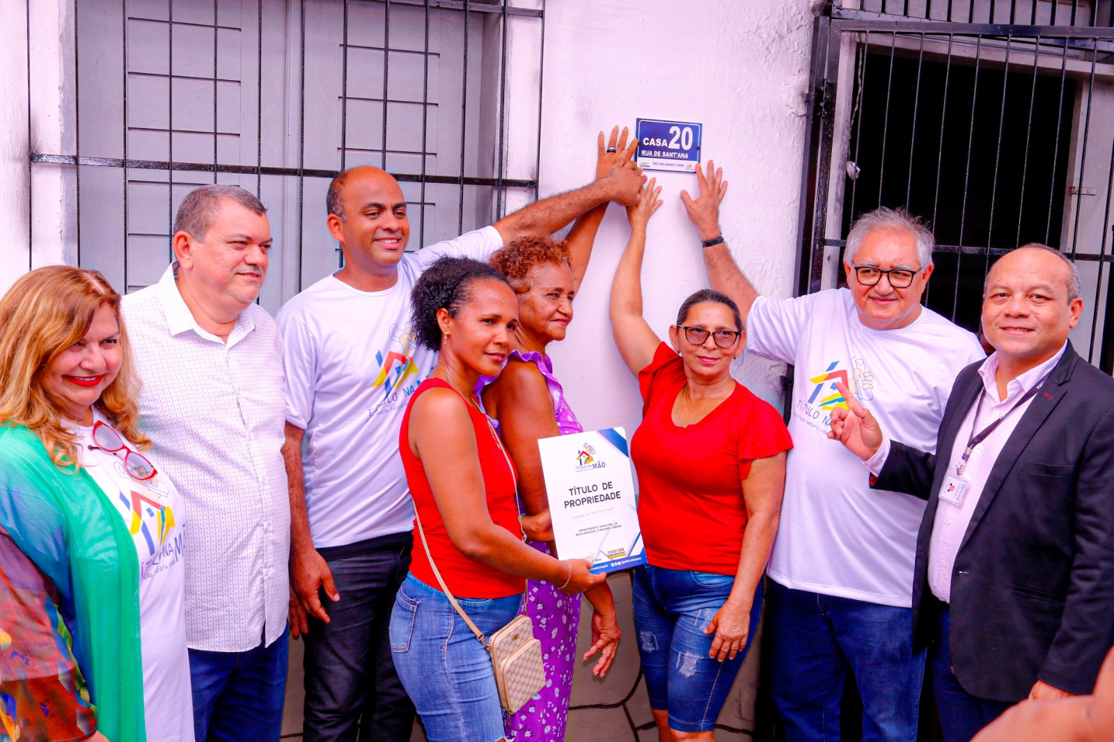 HOMENS E MULHERES DE PÉ EM FRENTE A FACHADDA DE CASA, APONTANDO PARA PLACA AZUL COM NÚMERO 10. AO CENTRO DUAS MULHERES VESTIDAS DE VERMELHO SEGURAM DOCUMENTO.