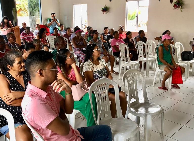 FOTO HORIZONTAL DE HOMENS E MULHERES SENTADOS EM CADEIRAS BRANCAS  AUDITÓRIO DE PAREDES CLARAS, VISTOS DE LADO. AO FUNDO, DO LADO DIREITO, DUAS JANELAS. DO LADO ESQUERDO, UMA PORTA DE VIDRO.