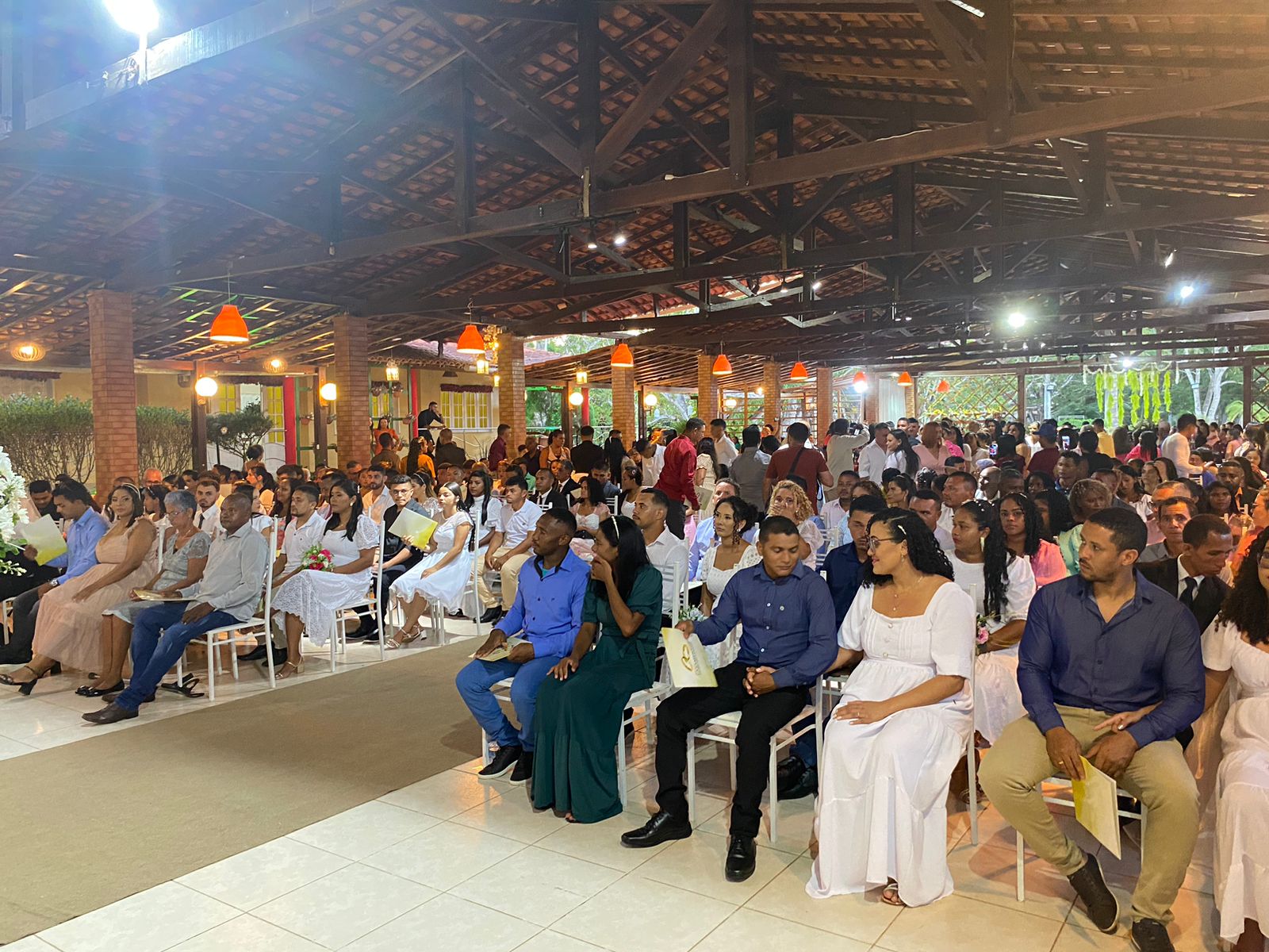 FOTO HORIZONTAL, COLORIDA, DE HOMENS E MULHERES EM CERIMÔNIA DE CASAMENTO COMUNITÁRIO,  SENTADAOS EM FILIERAS DE CADEIRAS BRANCAS EM SALÃO, COM TETO DE TELHA E MADEIRA, TAPETE BEGE AO CENTRO.