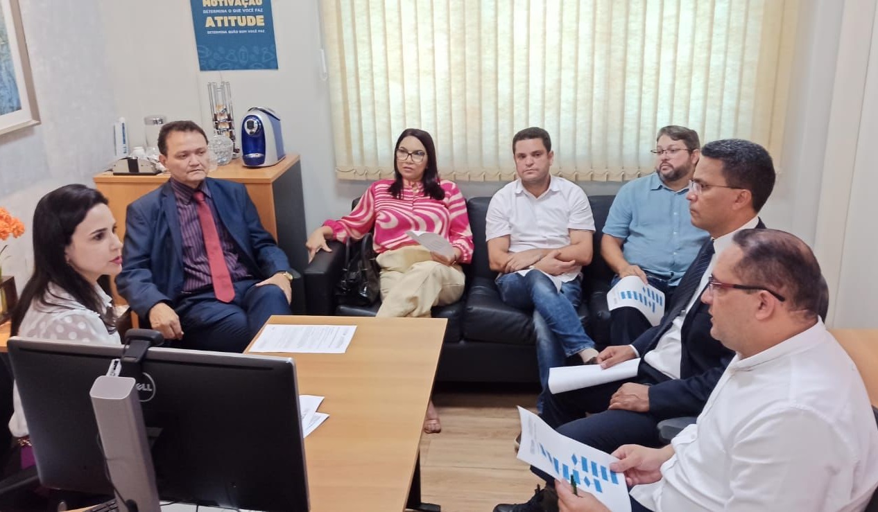 FOTO HORIZONTAL, COLORIDA, DE HOMENS E MULHERES EM REUNIÃO, EM SALA DE PAREDES CLARAS, EM TORNO DE MESA DE MADEIRA. AO FUNDO, CORTINA CLARA. EM PRIMEIRO PLANO, À ESQUERDA, MONITOR DE TV VISTO POR TRÁS.