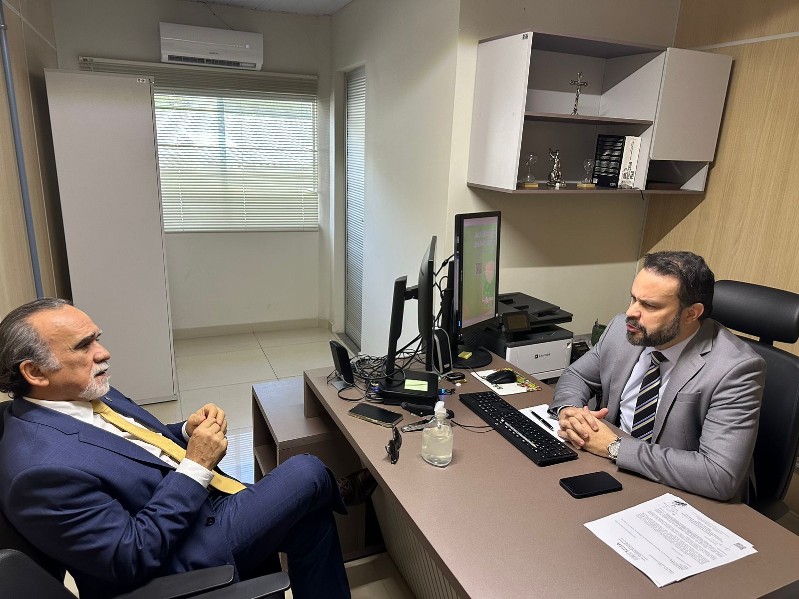 Foto colorida. Em um ambiente fechado - um gabinete - dois homens dialogam em lados opostos sentados em uma mesa.