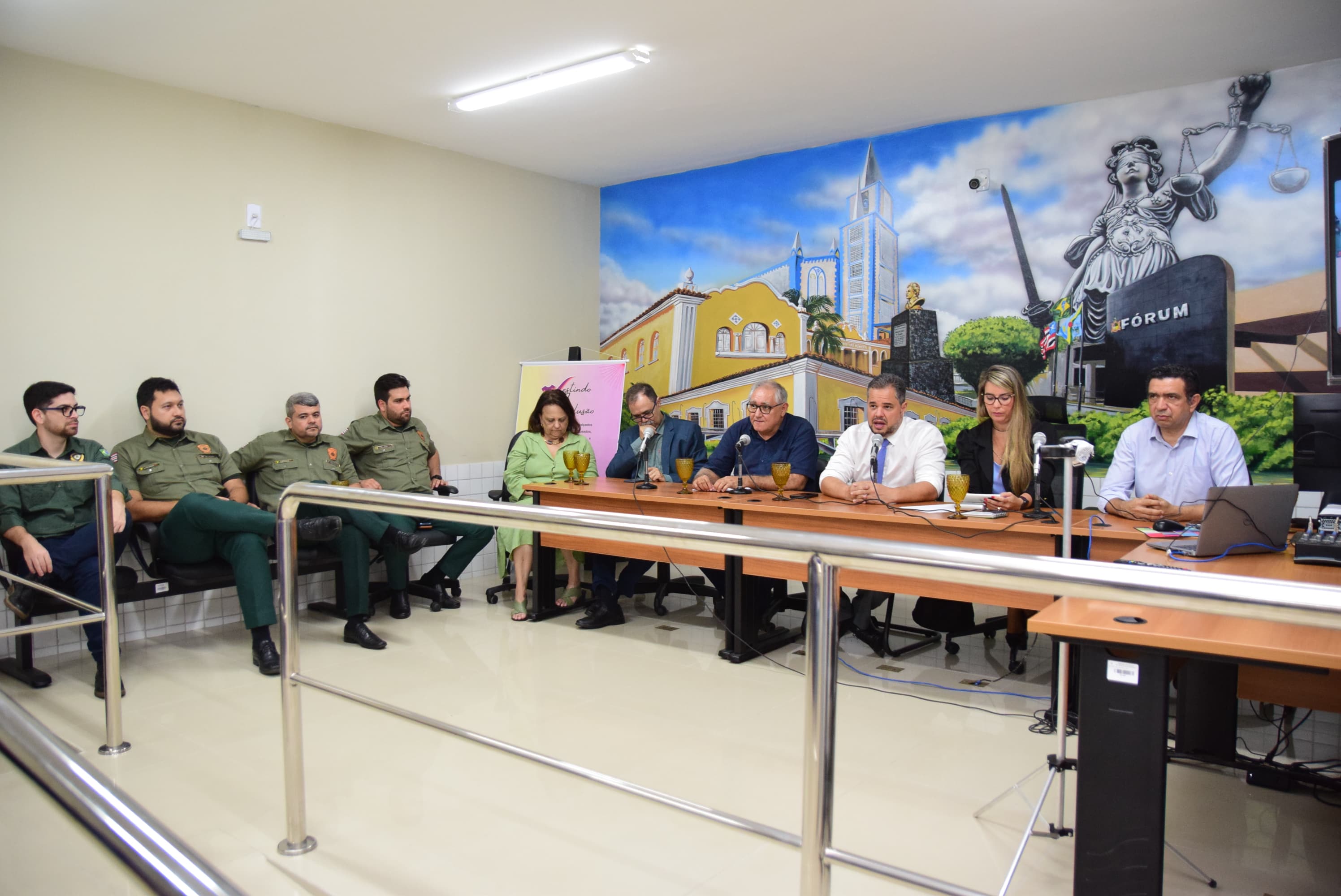 FOTO HORIZPNTAL, COLORIDA, DE HOMENS E MULHERES SNETADOS ATRÃS DE MESA, EM AUDITÓRIO COM PAREDE COM PINTURA COM TEMA DE JUSTIÇA AO FUNDO. EM PRIMEIRO PLANO, GRADIAL DE AÇO INOX.