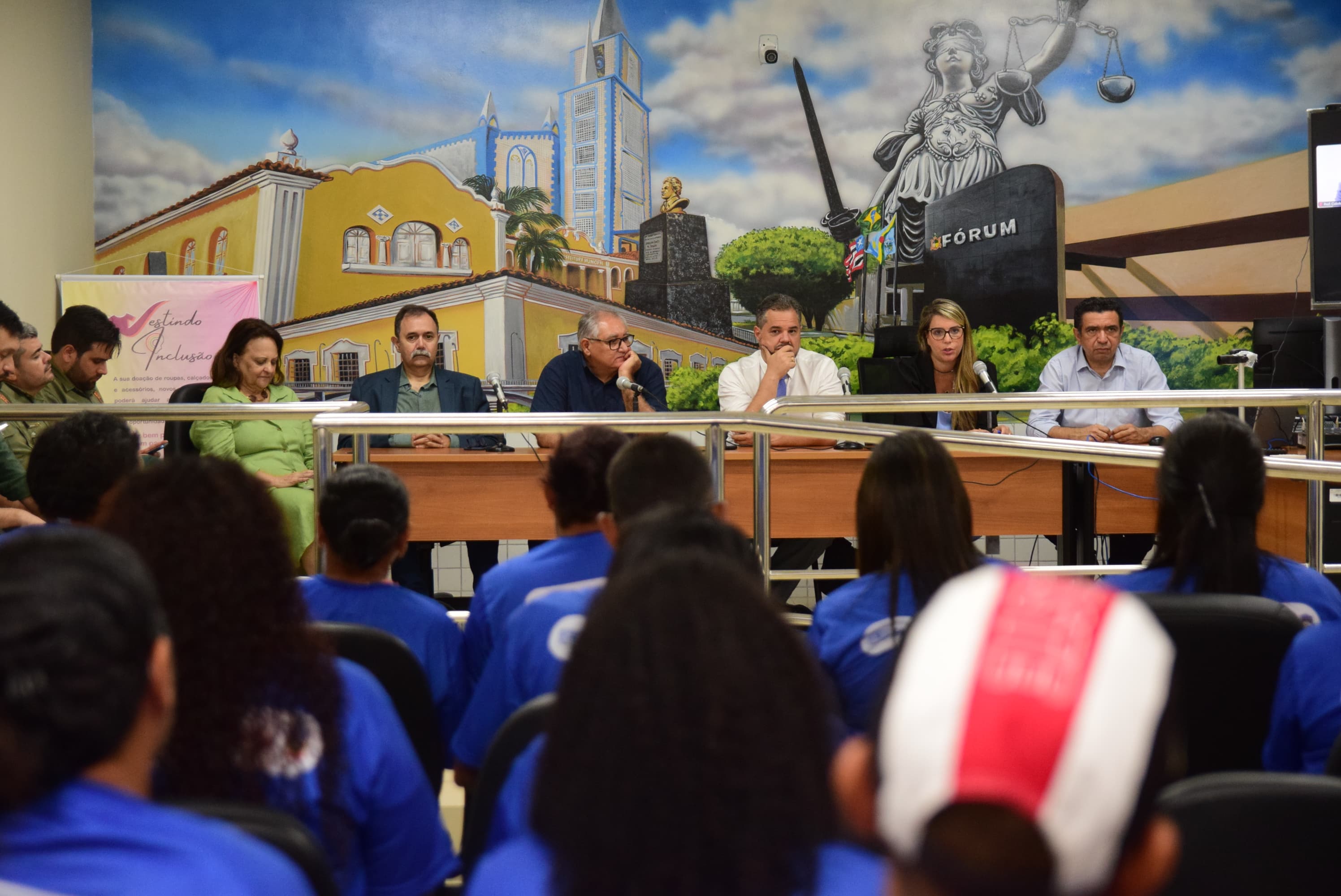 FOTO HORIZONTAL, COLORIDA, DE HOMENS E MULHERES VISTOS DE COSTAS, SENTADOS EM AUDITÓRIO, VESTIDAS DE CAMISETAS AZUIS. AO FUNDO, HOMENS E MULHERES SENTADOS ATRÁS DE MESA DIANTE DE PAREDE COM PINTURA COM TEMA DE JUSTIÇA.