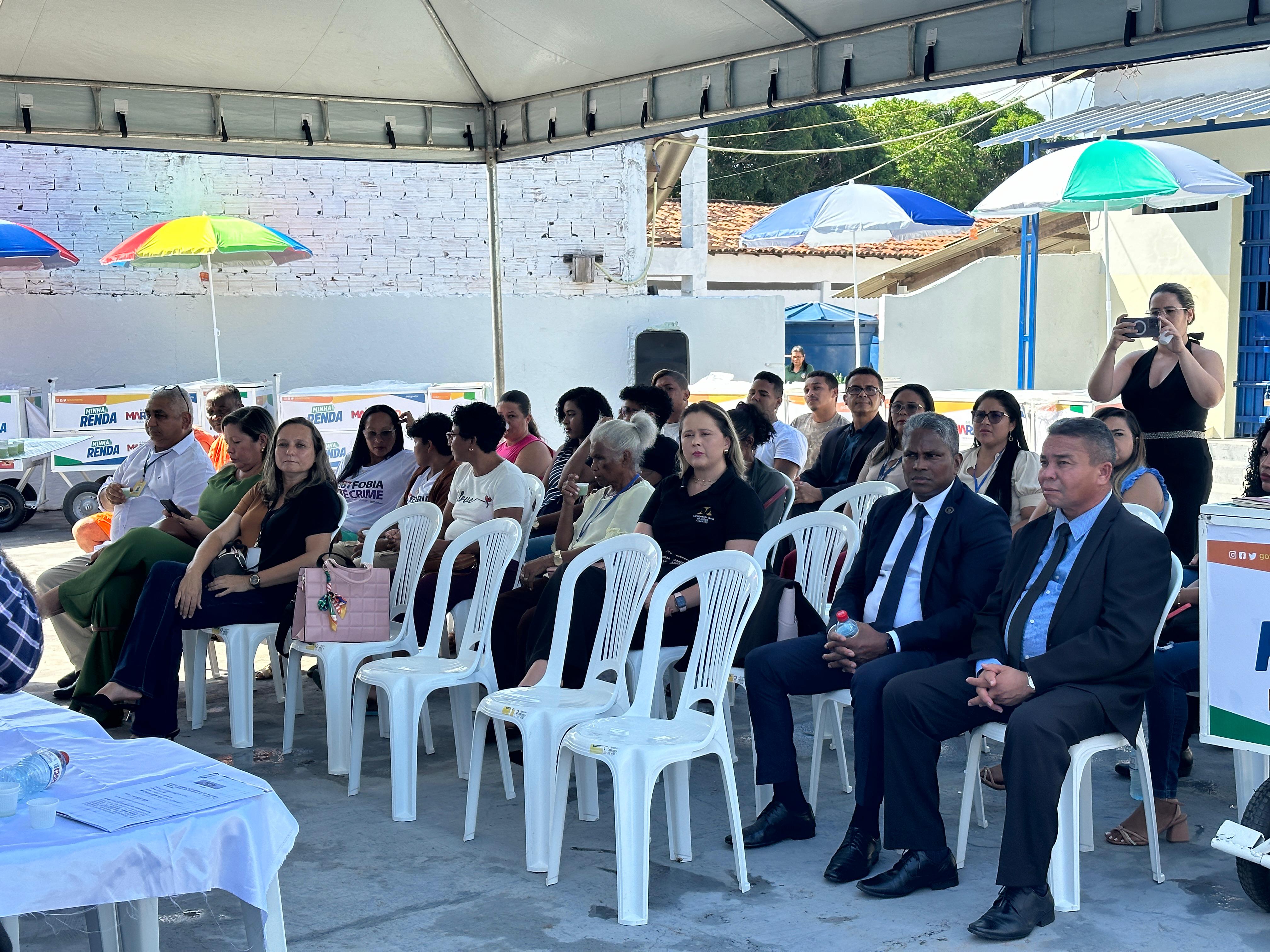 A imagem mostra um grupo de pessoas sentadas em uma longa mesa com um pano branco, em frente a uma parede com vários cartazes. Os rostos das pessoas estão obscurecidos por blocos retangulares por razões de privacidade. A mesa tem microfones e papéis, sugerindo que isso pode ser um painel de discussão ou reunião formal. Há também uma planta à esquerda, no primeiro plano. A parede atrás apresenta cartazes com texto e imagens, indicando que esse evento pode estar relacionado a atividades culturais ou educacionais. Um dos cartazes menciona ¿DIÁLOGOS RESTAURATIVOS¿, outro faz referência a um simpósio internacional sobre direitos humanos, e outros parecem anunciar eventos artísticos ou literários.