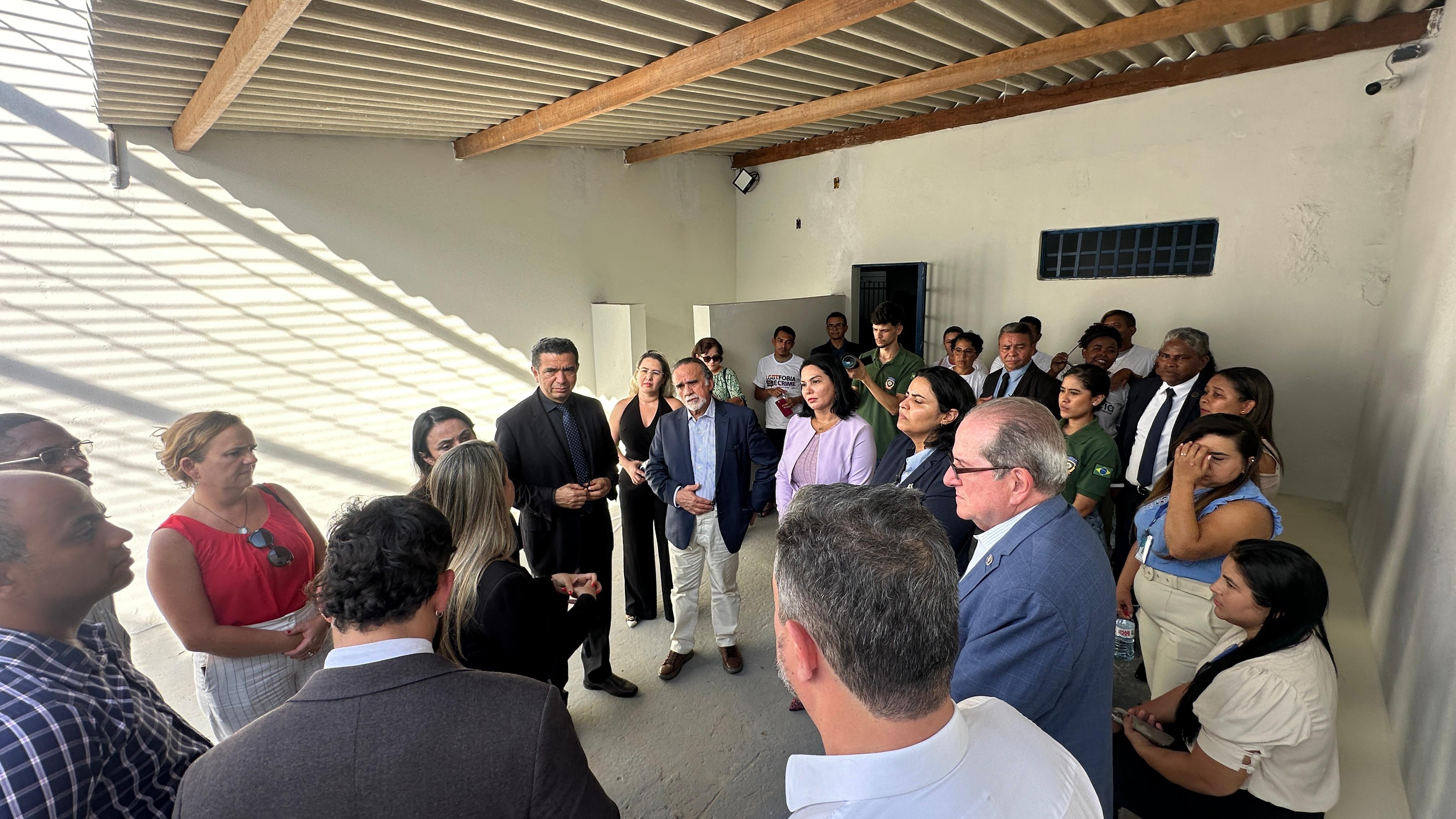 A imagem mostra um grupo de pessoas sentadas em uma longa mesa com um pano branco, em frente a uma parede com vários cartazes. Os rostos das pessoas estão obscurecidos por blocos retangulares por razões de privacidade. A mesa tem microfones e papéis, sugerindo que isso pode ser um painel de discussão ou reunião formal. Há também uma planta à esquerda, no primeiro plano. A parede atrás apresenta cartazes com texto e imagens, indicando que esse evento pode estar relacionado a atividades culturais ou educacionais. Um dos cartazes menciona ¿DIÁLOGOS RESTAURATIVOS¿, outro faz referência a um simpósio internacional sobre direitos humanos, e outros parecem anunciar eventos artísticos ou literários.