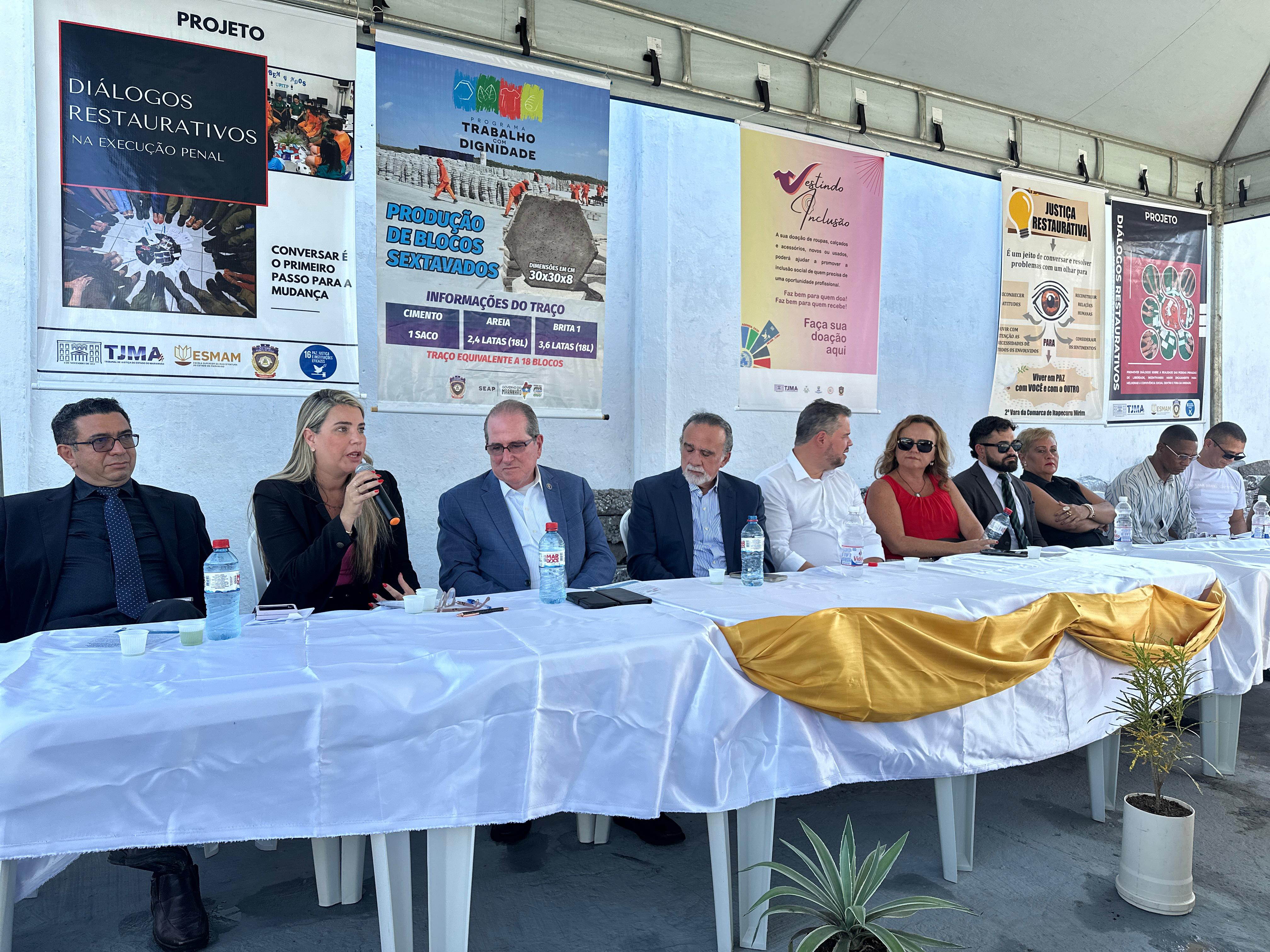 A imagem mostra um grupo de pessoas sentadas em uma longa mesa com um pano branco, em frente a uma parede com vários cartazes. Os rostos das pessoas estão obscurecidos por blocos retangulares por razões de privacidade. A mesa tem microfones e papéis, sugerindo que isso pode ser um painel de discussão ou reunião formal. Há também uma planta à esquerda, no primeiro plano. A parede atrás apresenta cartazes com texto e imagens, indicando que esse evento pode estar relacionado a atividades culturais ou educacionais. Um dos cartazes menciona ¿DIÁLOGOS RESTAURATIVOS¿, outro faz referência a um simpósio internacional sobre direitos humanos, e outros parecem anunciar eventos artísticos ou literários.