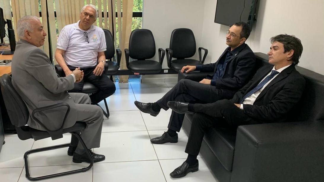 FOTO HORIZONTAL, COLORIDA, DE QUATRO HOMENS RM REUNIÃO EM SALA DE PISO BRANCO. À DIREITA, DOIS DE TERNOS ESCUROS SENTADDOS EM SOFÁ PRETO. À ESQUERDA, OUTROS DOIS DE ROUPA CLARA, SENTADOS EM CADEIRAS.AO FUNDO, CORTINA AMARELA CLARA E FILEIRA DE CADEIRAS PRETAS.