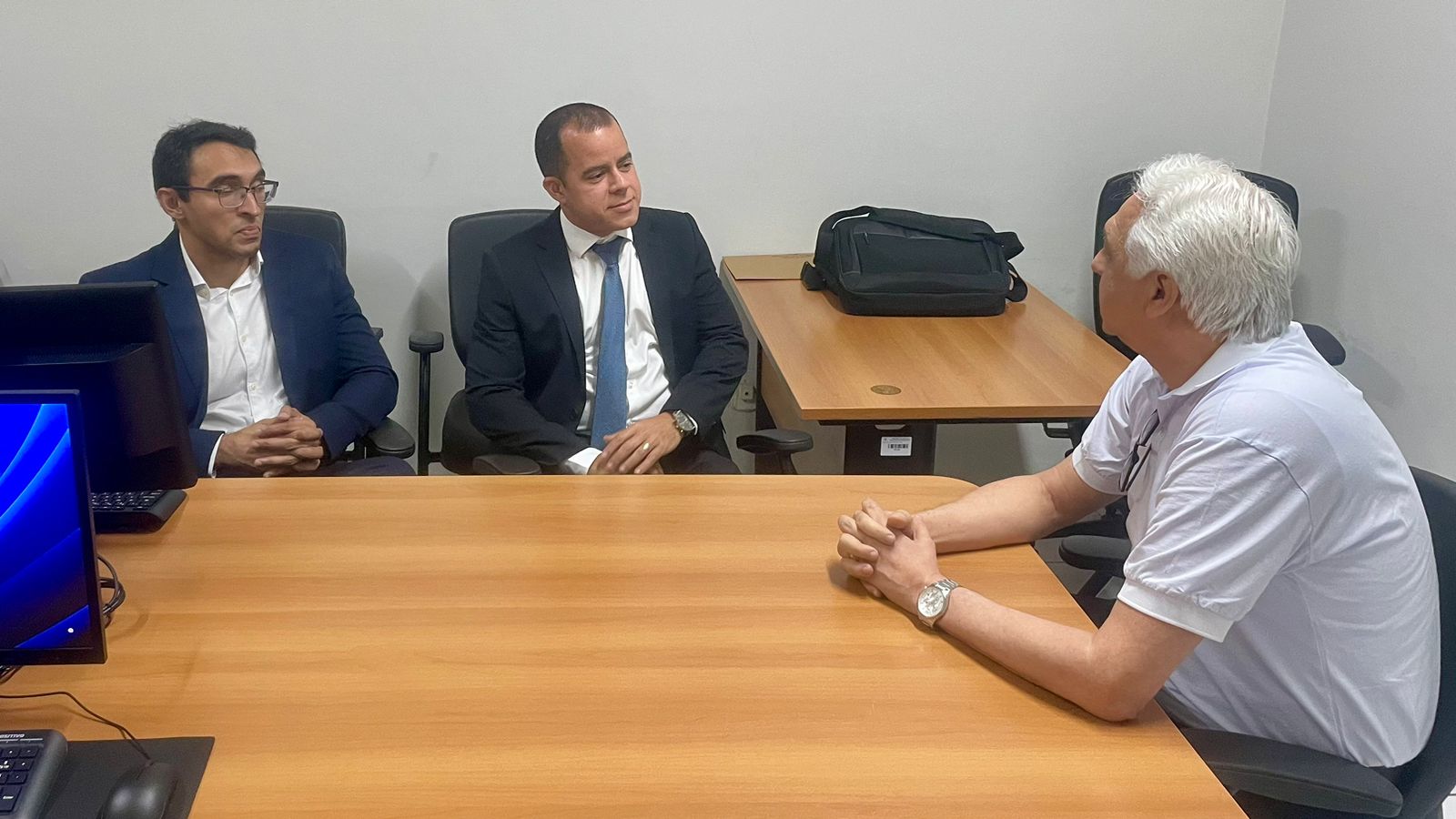 Foto horizontal, colorida, de três homens conversando em torno de mesa de madeira clara, em sala de trabalho. À esquerda, homem pardo, de terno escuro com uma camisa branca. Ao centro, homem branco, de terno escuro e camisa branca,  com uma gravata escura.
À direita, homem branco, cabelos brancos, de camisa cinza claro com as mangas dobradas e braços sobre a mesa. No direito, um relógio no pulso esquerdo.