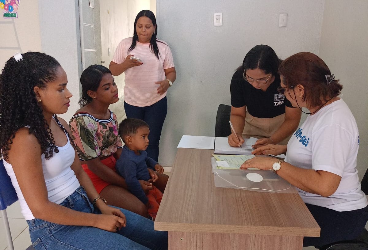 Imagem horizontal, colorida, de seis pessoas em torno de uma mesa de madeira, em  ambiente interno, cinco sentadas e uma de pé. À esquerda, mulher usando uma blusa preta sem mangas com alças e calça jeans azul. Ao lado,  outra veste blusa rosa de mangas curtas e calças escuras, com um menino no colo, usando uma camisa azul com padrões brancos e short vermelho. Ao centro, mulher de pé, vestindo uma blusa de cor clara com mangas dobradas e calças escuras. 
Na extremidade direita da mesa, alguém está sentado, usando uma camisa branca de mangas curtas com algum texto ou logotipo e calças claras.