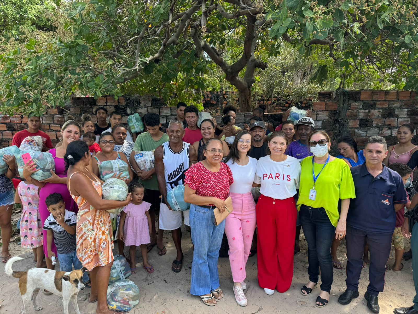 Foto horizontal colorida, de um grupo de pessoas em pé, em área livre. Ao fundo, área verde e uma parede de tijolos.
Há também um cão branco no canto inferior esquerdo da imagem