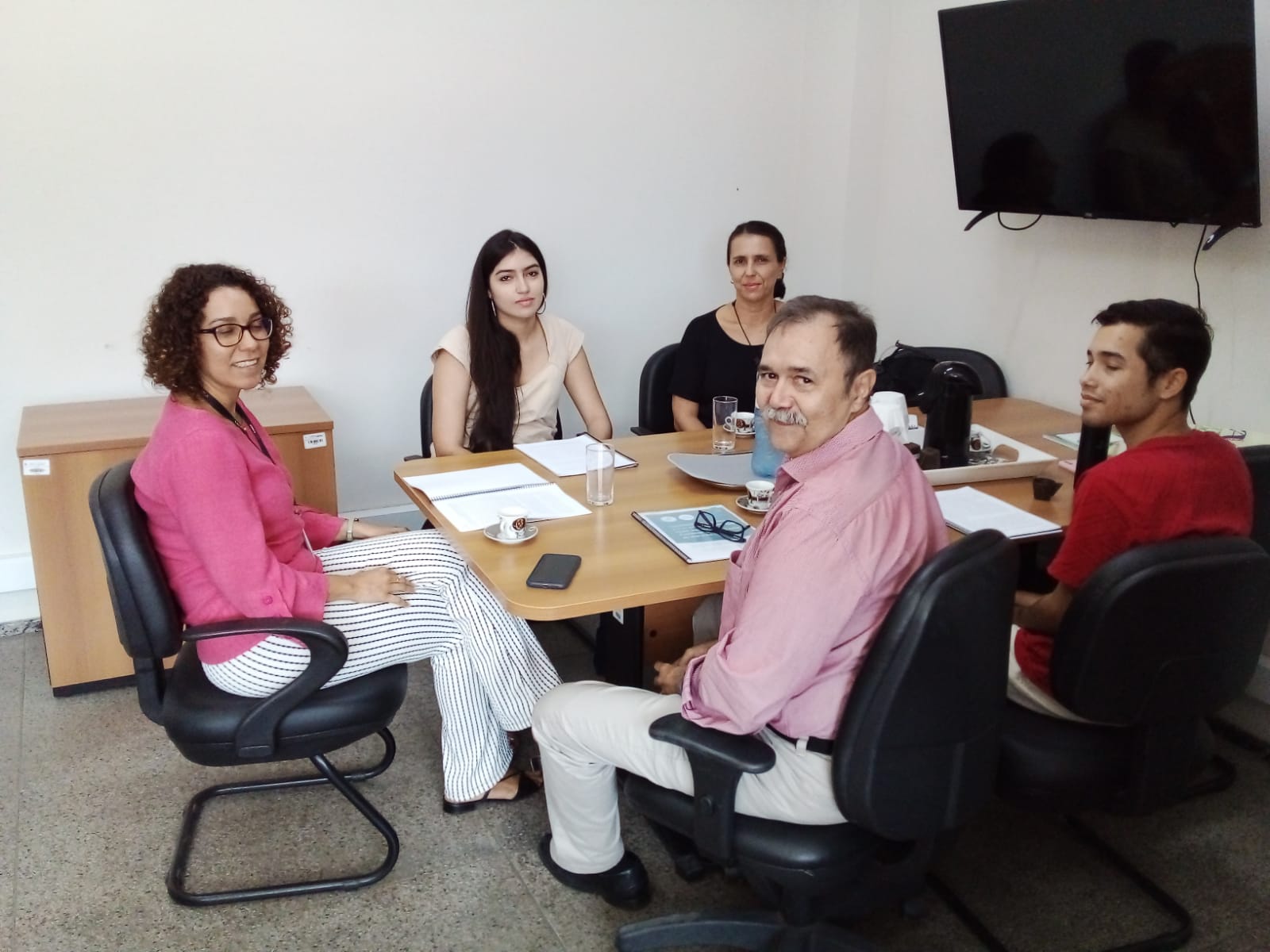 Foto horizontal, colorida, de três mulheres e dois homens em sala de trabalho, sentados em torno de mesa. À direita, um monitor de TV está fixado em parede branca. Na mesa, há papéis, um laptop e alguns utensílios de escrita.