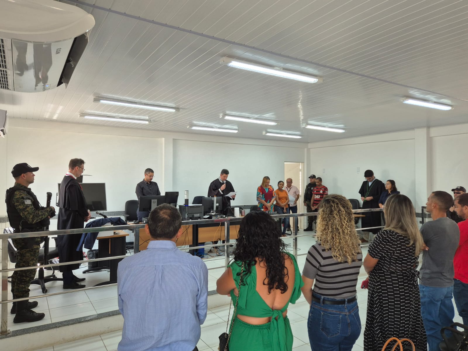 Foto horizontal, colorida, de sala de tribunal do júri. Há várias pessoas em pé e algumas sentadas. No primeiro plano, um grupo de pessoas está voltado para o centro da sala, onde três pessoas estão de costas para a câmera. No plano intermediário, uma pessoa vestindo uma toga preta, está de pé, atrás de uma mesa com microfones e papéis. À esquerda e à direita, há pessoas sentadas Ao fundo, há duas bandeiras em suportes e outra pessoa em traje militar ao lado delas.