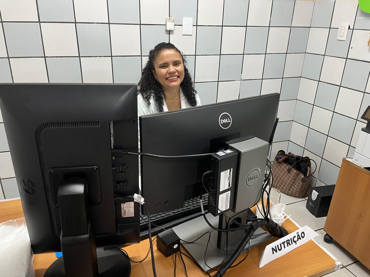 UMA MULHER ESTÁ ATRÁS DE DOIS COMPUTADORES SORRINDO EM UMA SALA. NA MESA ESTÁ A PLACA ESCRITO "NUTRIÇÃO"