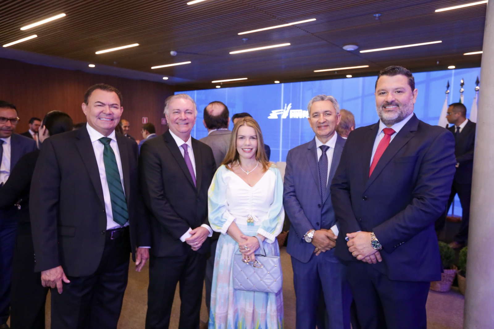 Foto horizontal, colorida, de um grupo de seis homens e uma mulher em pé lado a lado, em ambiente interno, com iluminação artificial e paredes decoradas com listras horizontais de madeira. Ao fundo, há uma parede iluminada com um logotipo corporativo e várias bandeiras penduradas verticalmente. Os indivíduos estão vestidos formalmente; os homens usam ternos e gravatas, enquanto a mulher, ao centro, usa um vestido claro e segura uma bolsa pequena.