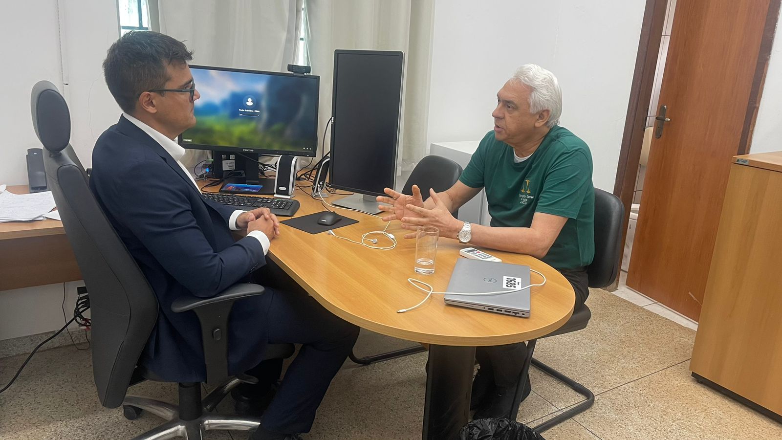ESTÃO SENTADOS Á MESA DOIS JUIZES. EM CIMA DA MESA HÁ UM COMPUTADOR COM DOIS MONITORES.
