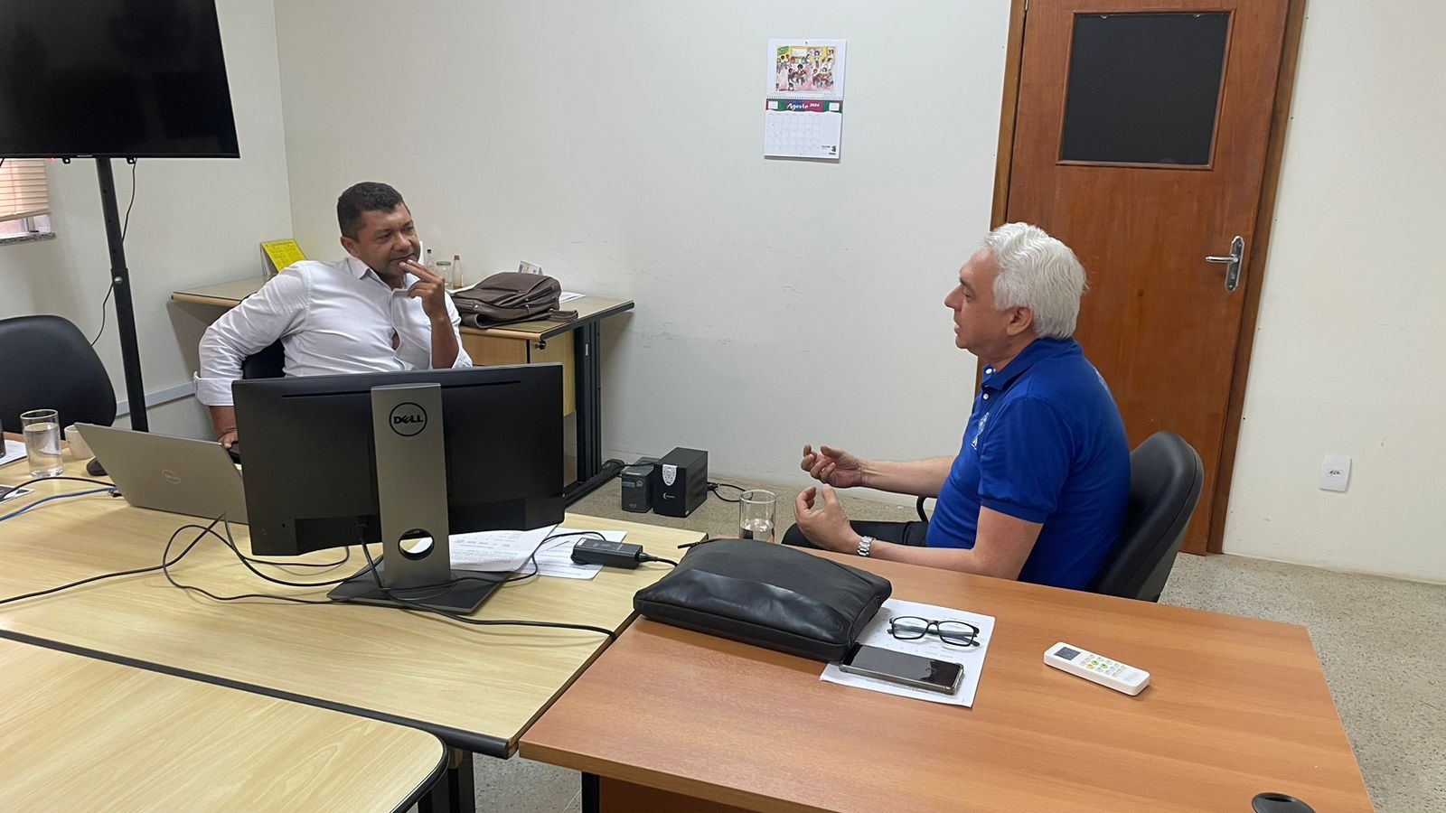 DOIS JUIZES EM REUNIÃO. NA FOTO HÁ DUAS MESAS JUNTAS. COM UM MONITOR. E DOIS COMPUTADORES. AO FUNDO, UMA PORTA DE MADEIRA ESCURA.