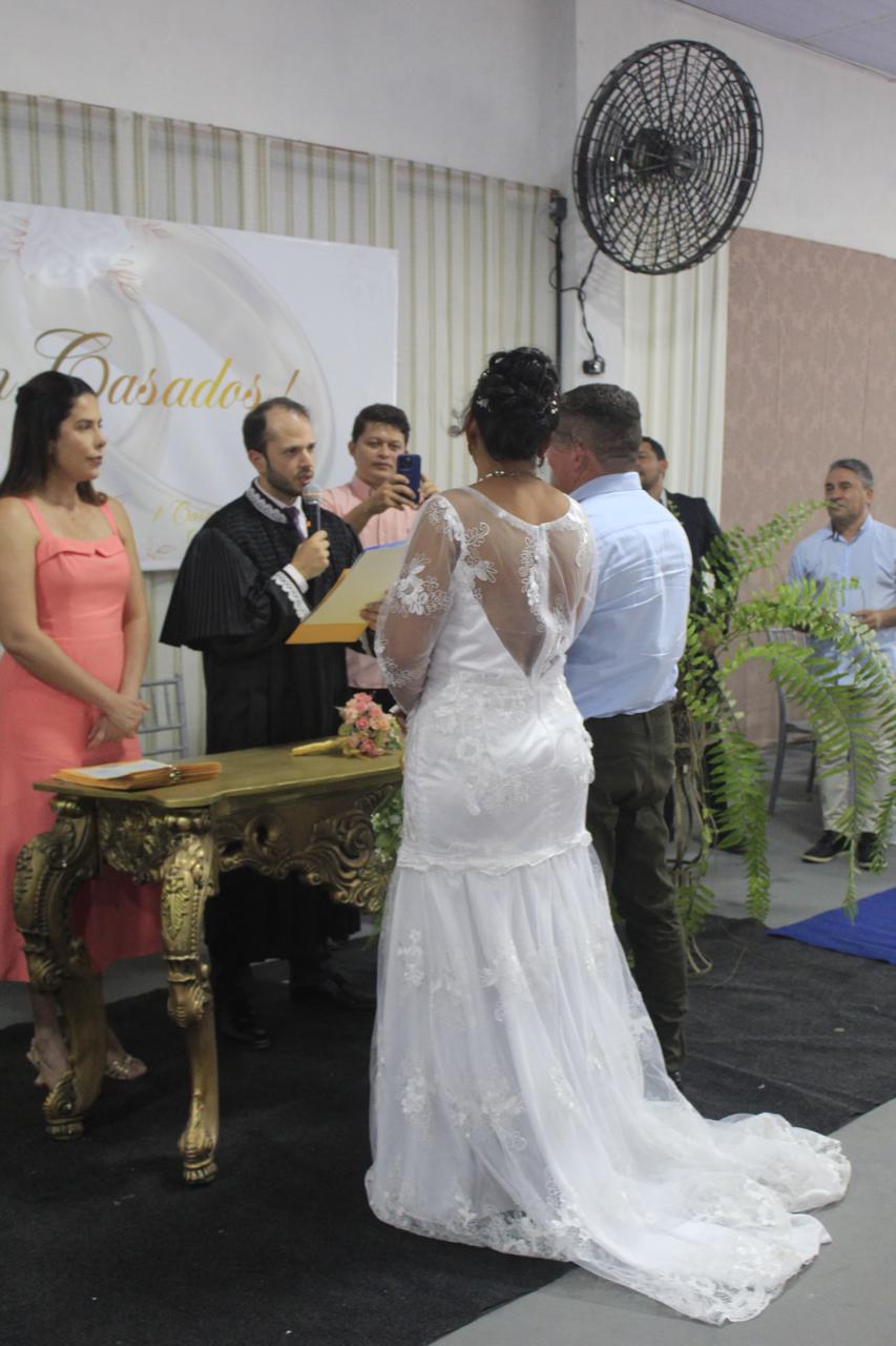 Foto vertical, colorida, de casamento, mostra casal de costas diante de um homem branco, de toga preta, ao lado de outras pessoas. A mulher usa vestido branco longo, de renda com mangas longas e costas abertas. O home, calça escura e camisa branca. Um cartaz na parede tem a palavra ¿Casados¿, ao fundo.