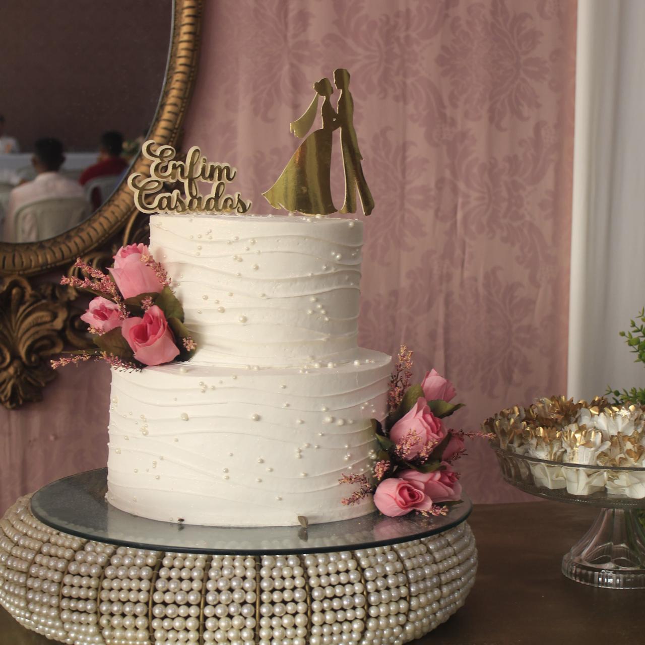 Foto horizontal, colorida, de bolo nupcial de dois andares, branco, adornado com flores na cor rosa clara. com topo de casal, sobre base redonda, com pérolas. Ao fundo, painel rosa com espelho dourado. À Direita, detalhe de louça com doces.