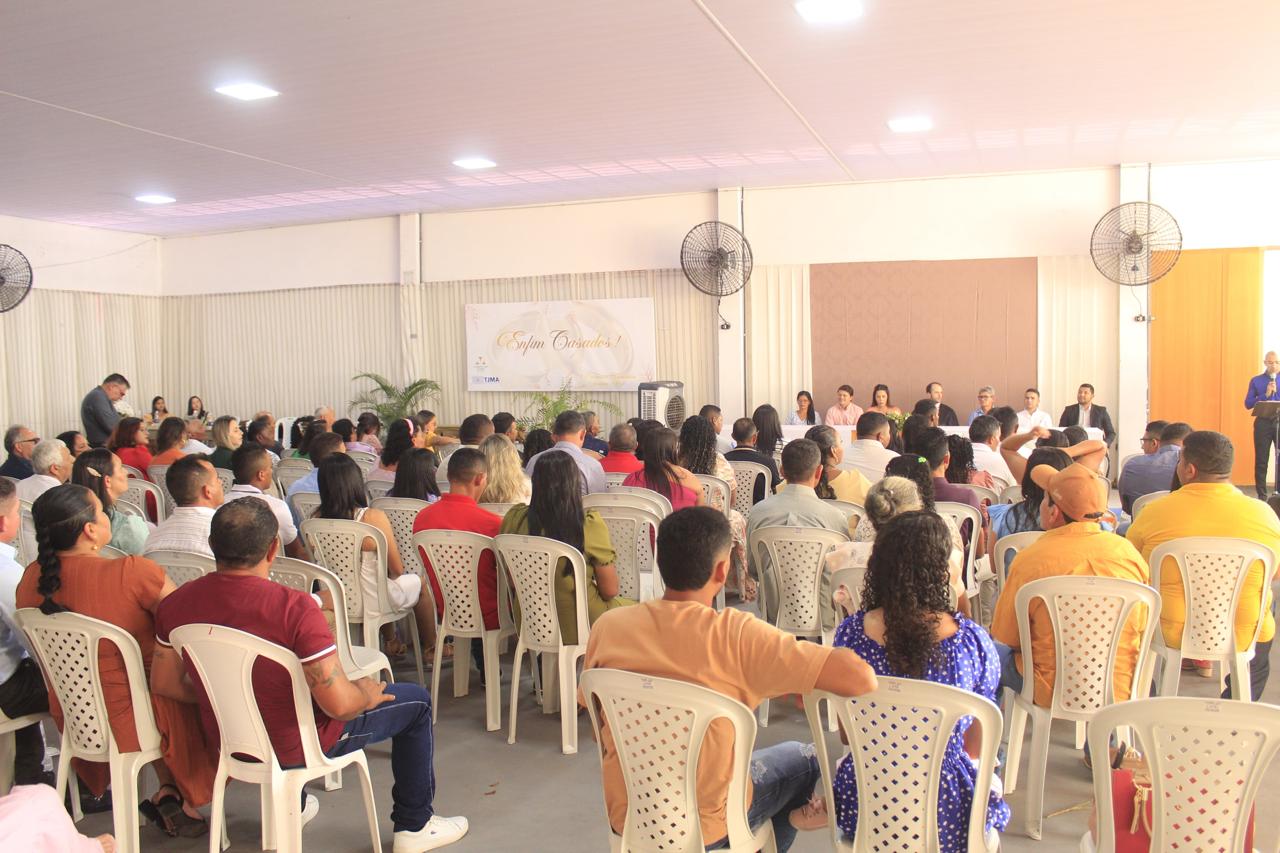 Foto horizontal, colorida, em um ambiente interno. Homens e mulheres estão sentados em cadeiras dispostas em fileiras, voltadas para um palco onde um grupo de indivíduos está sentado em uma mesa longa coberta com uma toalha branca. Acima da mesa, há um banner com a inscrição "Felicidade". Há um orador que está no pódio à esquerda. O local possui paredes em cor clara e ventiladores de teto.