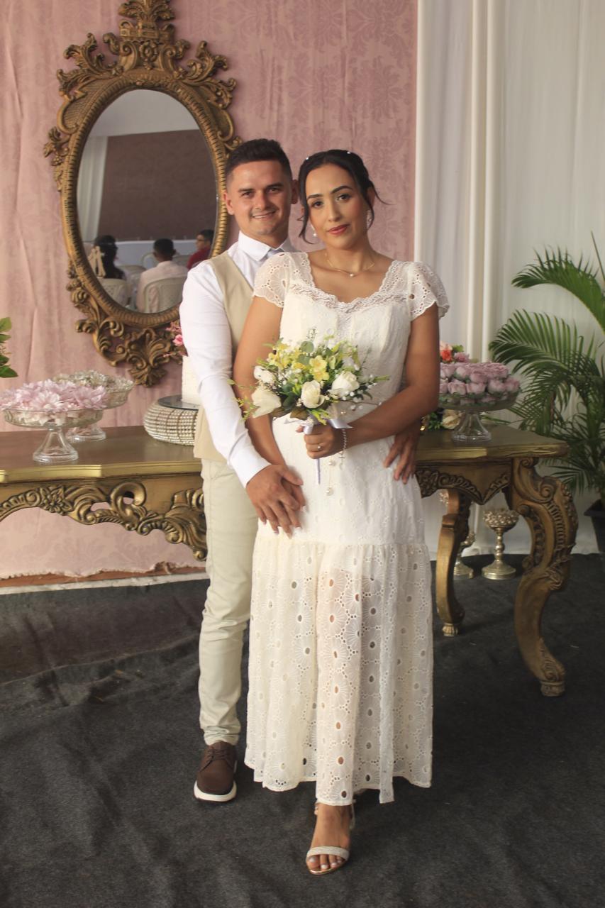 Foto vertical, colorida, de homem e mulher brancos, de pé, vestidos para casamento. Eles estão em frente um espelho dourado. A mulher usa vestido branco com detalhes em renda e segurando um buquê de flores. O homem está vestida com calças claras e uma camisa branca. Atrás deles, há um espelho dourado ornamentado refletindo suas imagens, e uma mesa com itens decorativos e flores.
