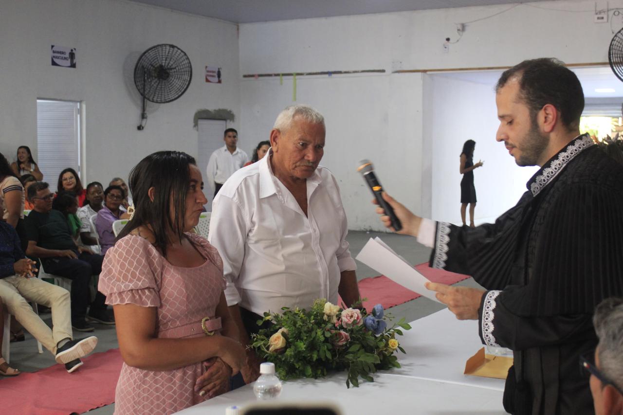 Foto horizontal, colorida, de casal de idosos diante de homem com toga preta de pé, com documento na mão esquerda e microfone na mão direita. Entre eles, há uma mesa coberta com toalha branca. O homem tem cabelos brancos e veste camisa branca. A mulher, parda, tem cabelos castanhos e compridos e usa vestido rosa. Ao fundo, pessoas sentadas em sala de paredes brancas.