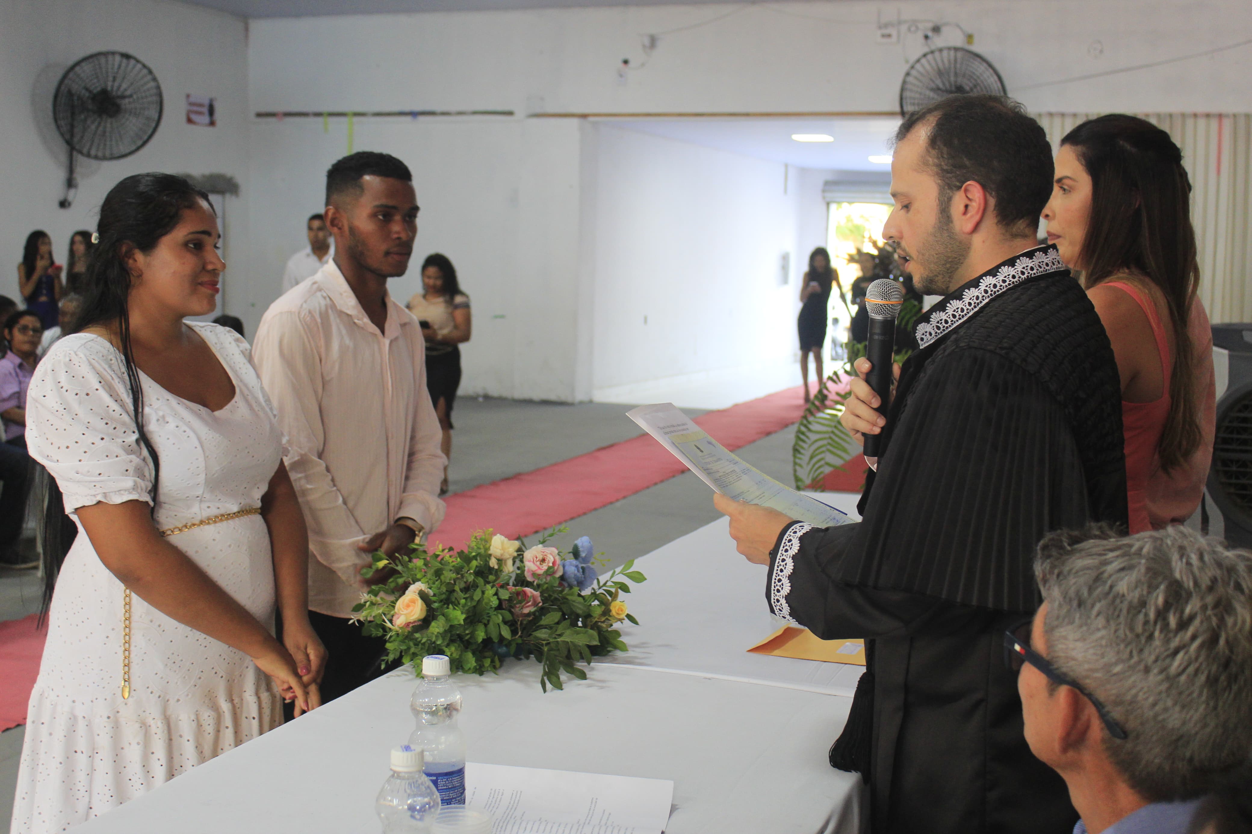 Foto horizontal, colorida, de casal de noivos pardos, de pé, vestidos de branco. Diante deles, um homem branco, de toga preta, segura documento com a mão esquerda e microfone com a mão direita. Entre eles há uma mesa coberta de toalha branca com arranjo verde com flores. Ao fundo, sala de paredes claras com tapete vermelho.