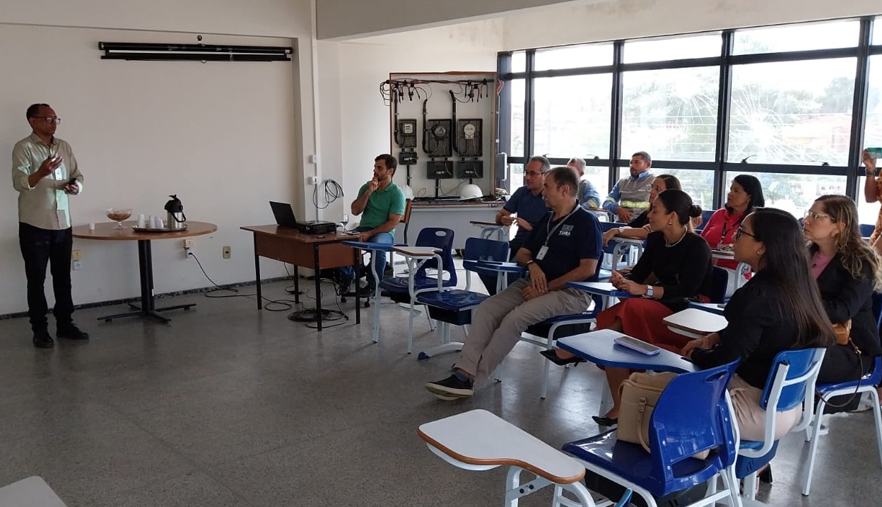 Foto horizontal, colorida, mostra uma sala com homens e mulheres sentadas em cadeiras azuis com pranchetas, voltadas para a esquerda da imagem onde um indivíduo está em pé apresentando algo. O apresentador está vestido com uma camisa clara e calça escura. À direita, há uma mesa com um laptop aberto e outros objetos. Ao fundo, há uma grande janela de vidro que permite a entrada de luz natural e mostra a vista exterior.