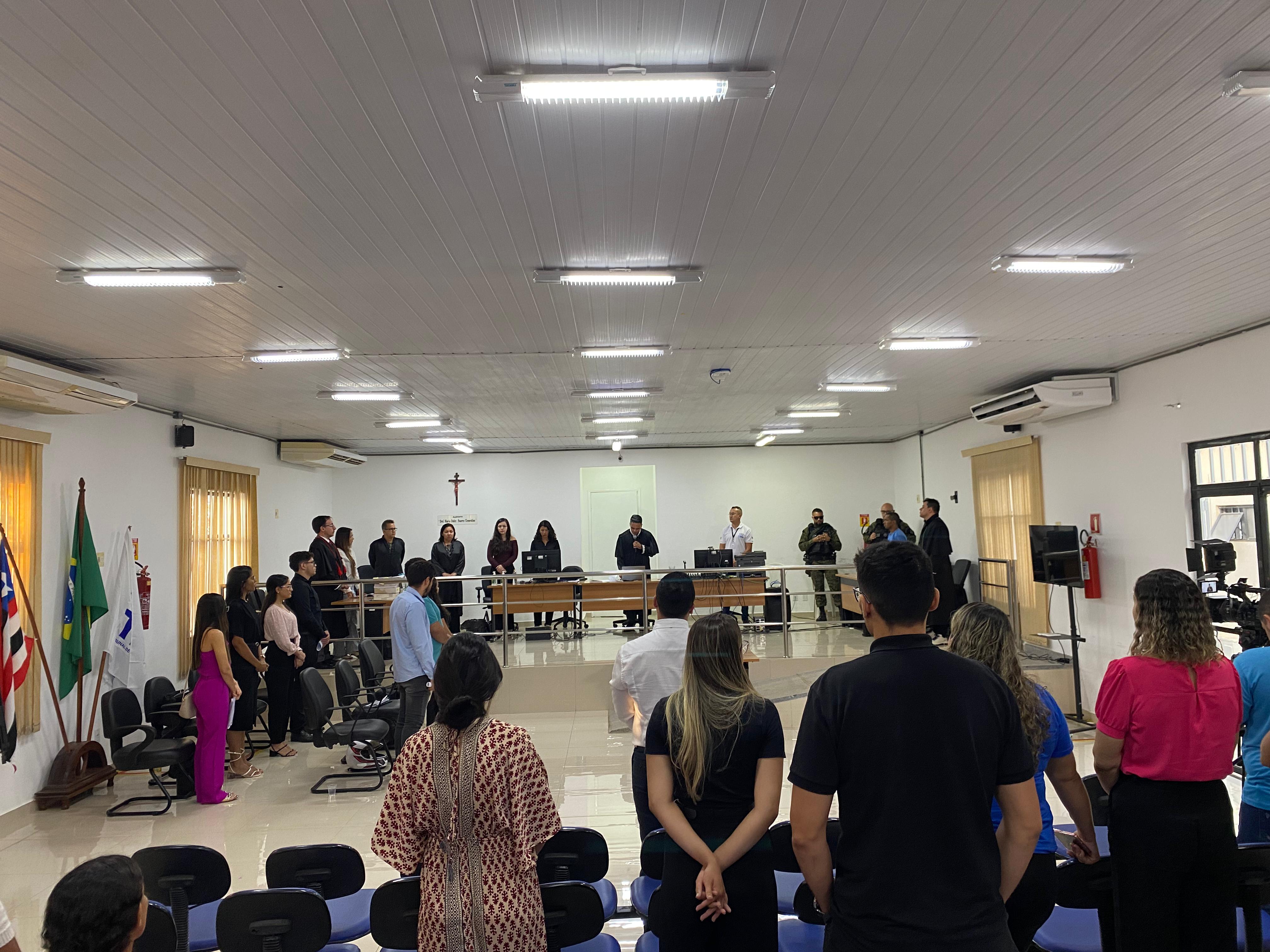 Foto horizontal, colorida, do salão do Tribunal do Júri, com pessoas vistas de costas, de pé, junto a cadeiras pretas, em primeiro plano. Ao fundo, mesa com computadores e, atrás dela,  homens de pé em frente a parede branca. Acima, teto branco, com três fileiras de luminárias acesas em perspectiva.