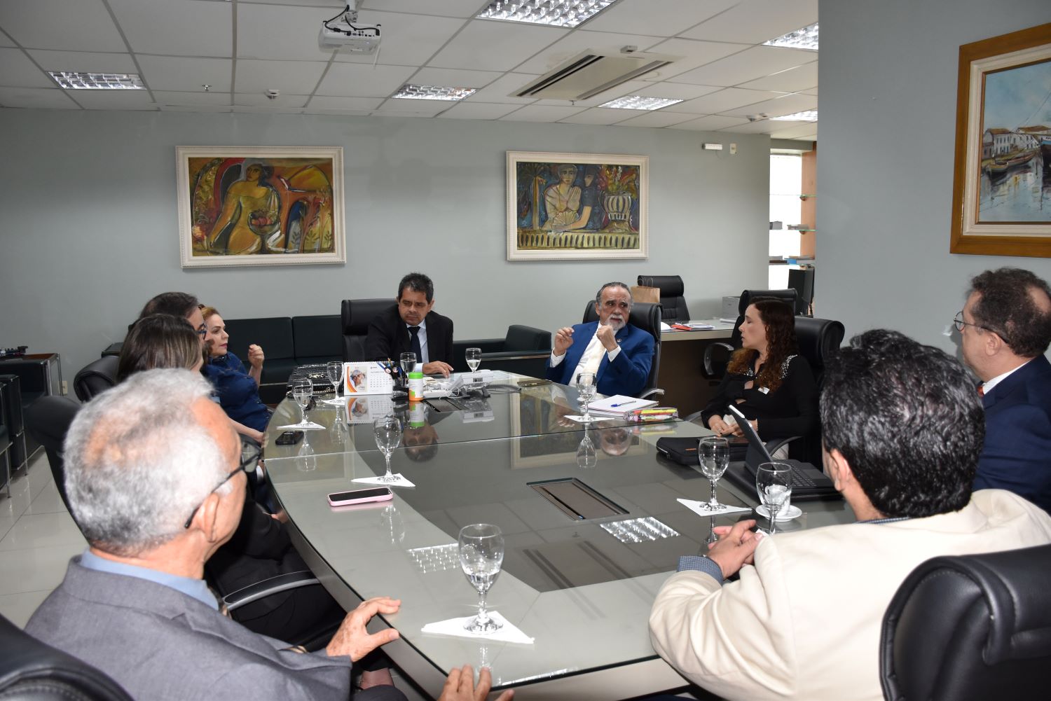 A imagem mostra uma reunião formal em uma sala de conferências. Sete pessoas estão sentadas ao redor de uma mesa de vidro retangular, onde há alguns copos de vidro com guardanapos, pastas e documentos sobre a mesa. As pessoas na reunião estão atentas e parecem discutir algum tema importante. Um dos homens ao centro, usando terno azul, gesticula com as mãos enquanto fala. Atrás dele, outro homem de terno preto observa a conversa.
