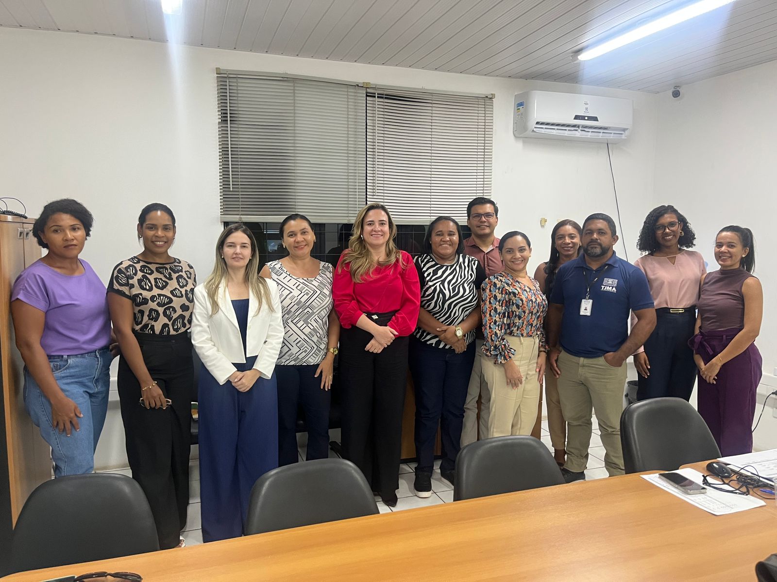 Foto horizontal, colorida, de mulheres de pé, posando para foto, em ambiente interno, com paredes brancas. Ao fundo, janela e aparelho de ar-condicionado. Em primeiro plano, uma mesa de madeira.